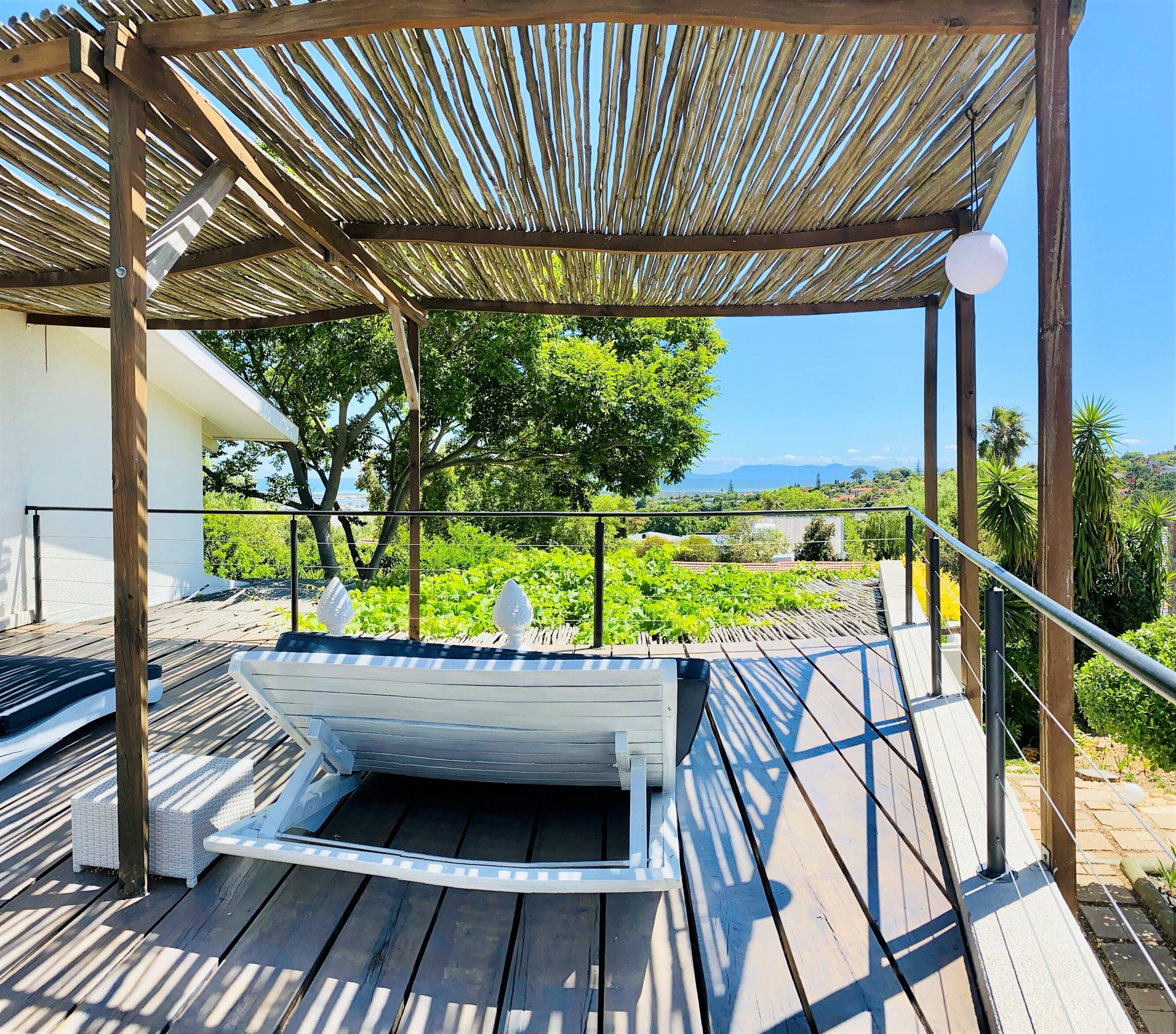 A wooden deck with a white boat-shaped hot tub, a pergola overhead, and a view of greenery and distant hills.