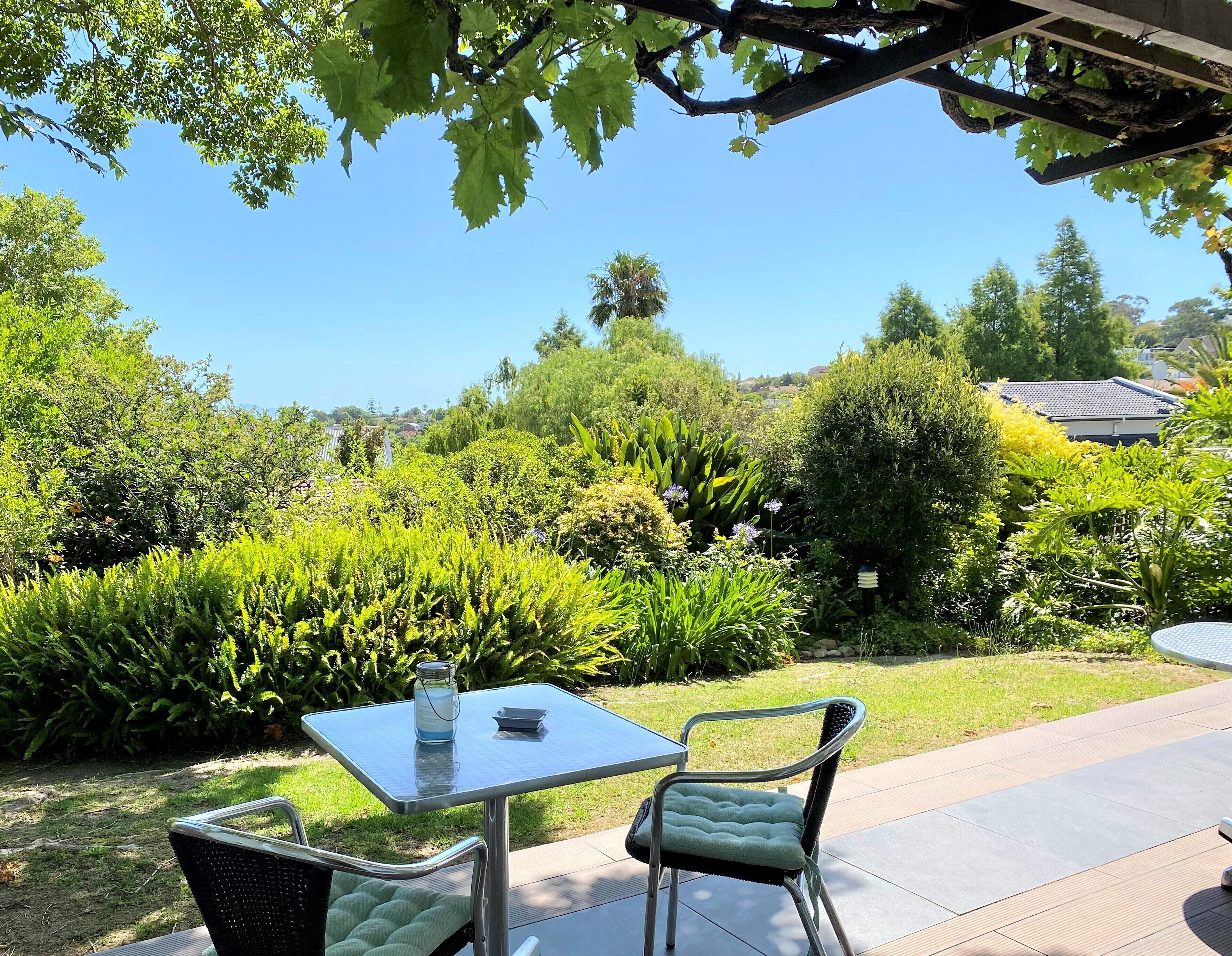 An outdoor patio with a table and chairs, surrounded by lush greenery and a clear blue sky.