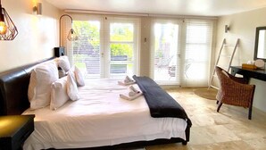 A bedroom with a large bed, a desk, a chair, and a view of the outdoors through the windows. - Southern Cross Guest House (Cape Town)