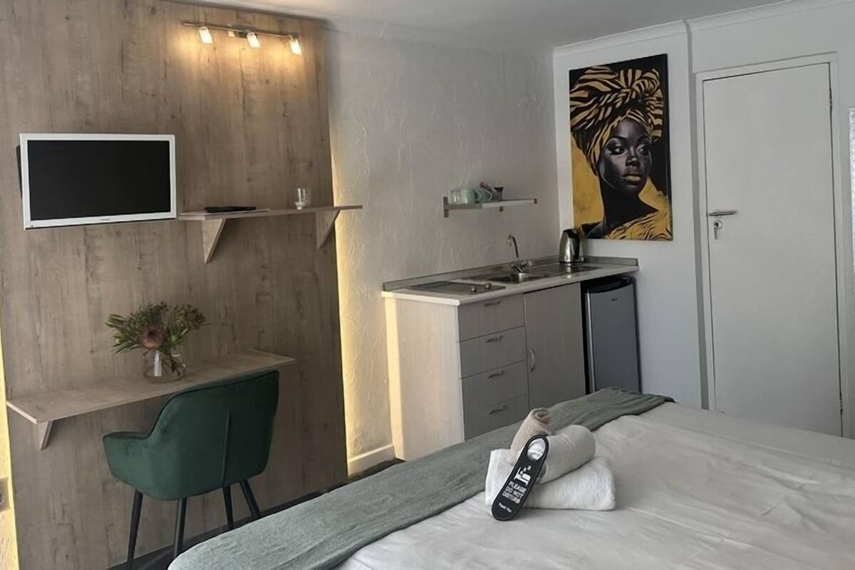A compact hotel room with a bed, a desk with a chair, a small kitchenette, and a mounted TV.