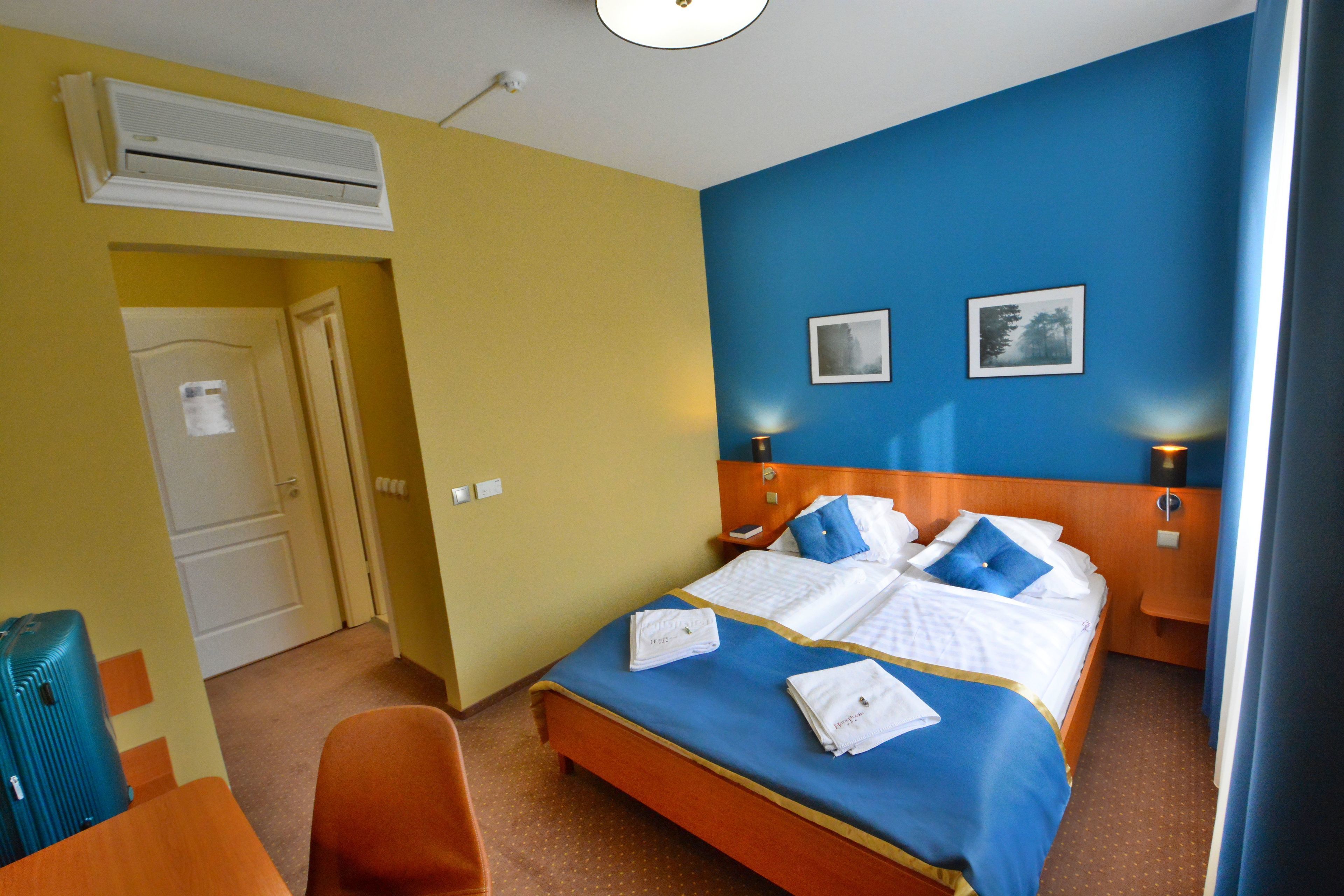 A hotel room with a bed, two bedside tables with lamps, a desk, a chair, an air conditioning unit, and a door.