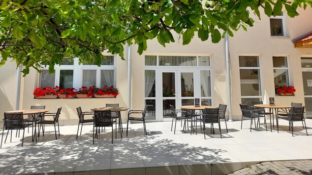 Eine Terrasse im Freien mit Tischen und Stühlen, umgeben von Grünpflanzen und einem Gebäude mit Fenstern.