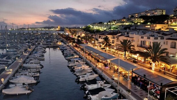 Un puerto deportivo con barcos amarrados a lo largo de un paseo marítimo, con edificios que ofrecen terrazas y colinas al fondo.