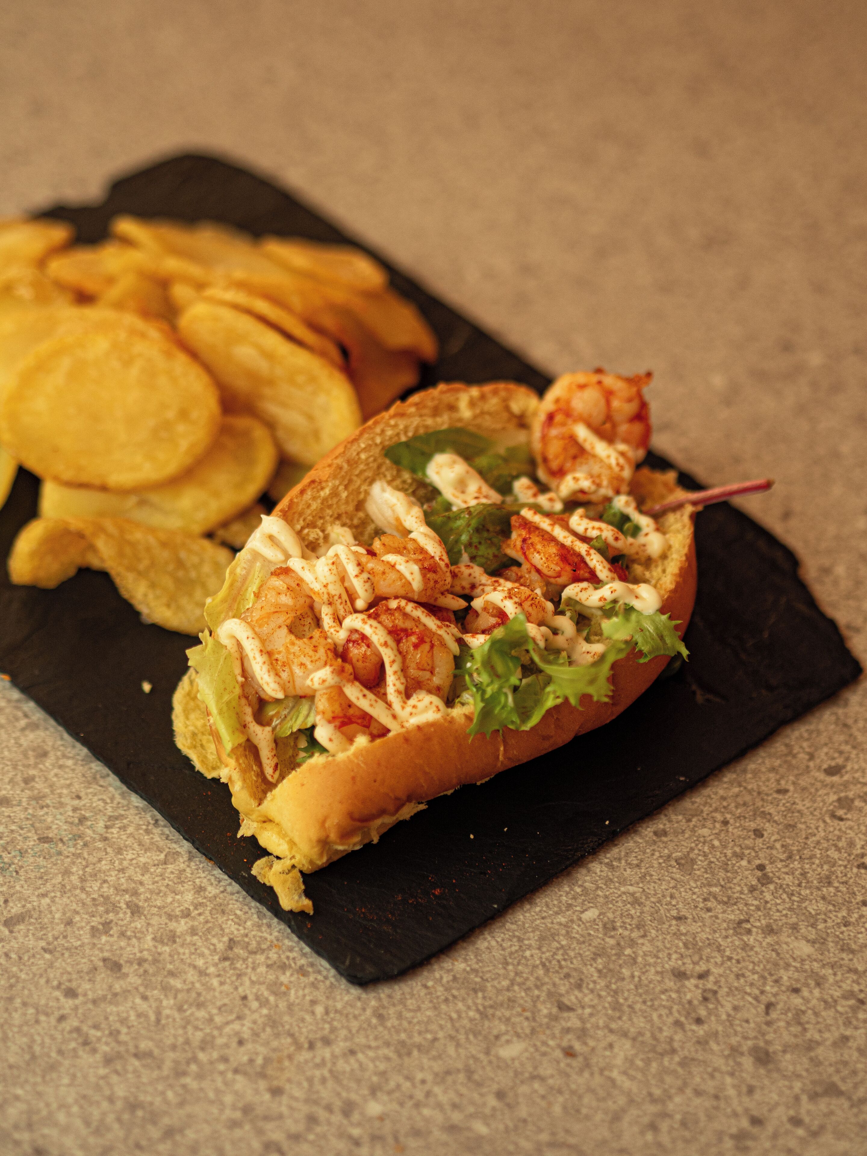 Un sándwich de camarones y lechuga sobre un plato negro, acompañado de papas fritas.