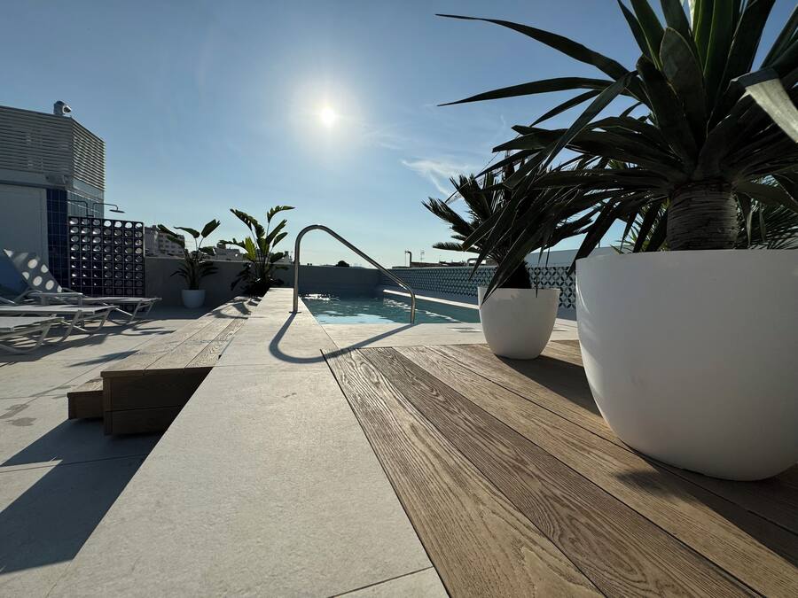 A rooftop pool area with lounge chairs, potted plants, and a clear view of the sea.