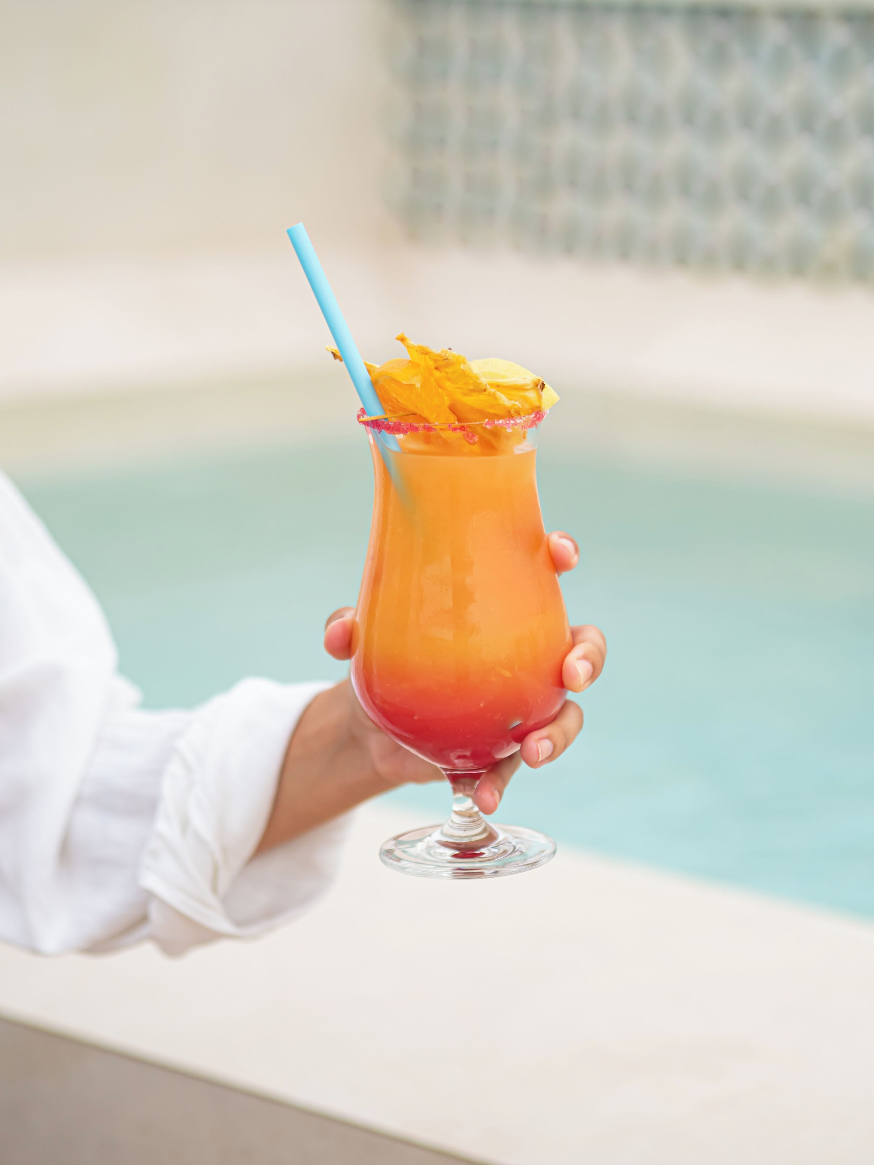 A person holding a cocktail glass with a blue straw and a tropical garnish, near a pool.