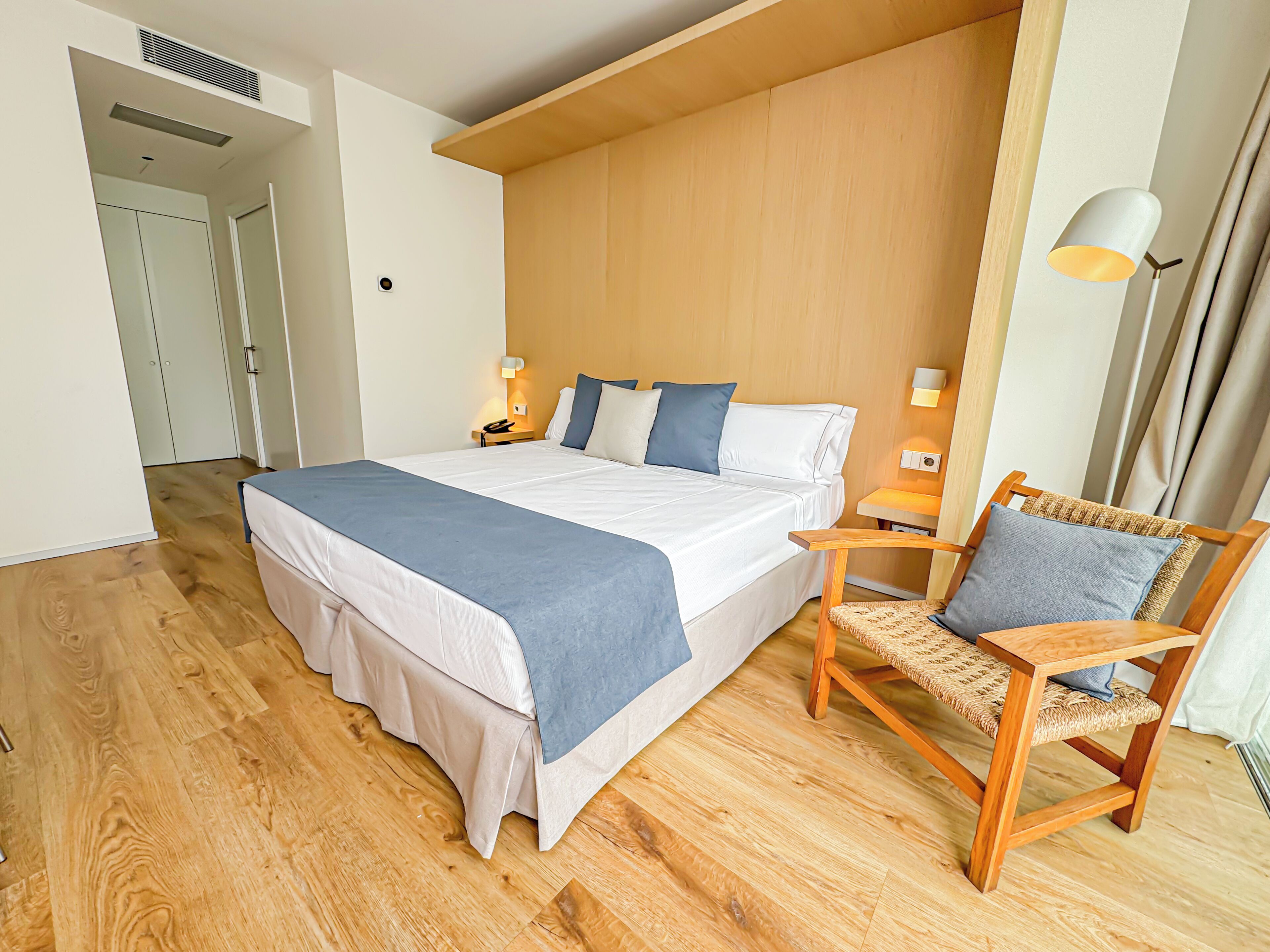 Une chambre d’hôtel moderne avec un grand lit, un plancher en bois, une chaise avec un siège en osier et une table de chevet avec une lampe.