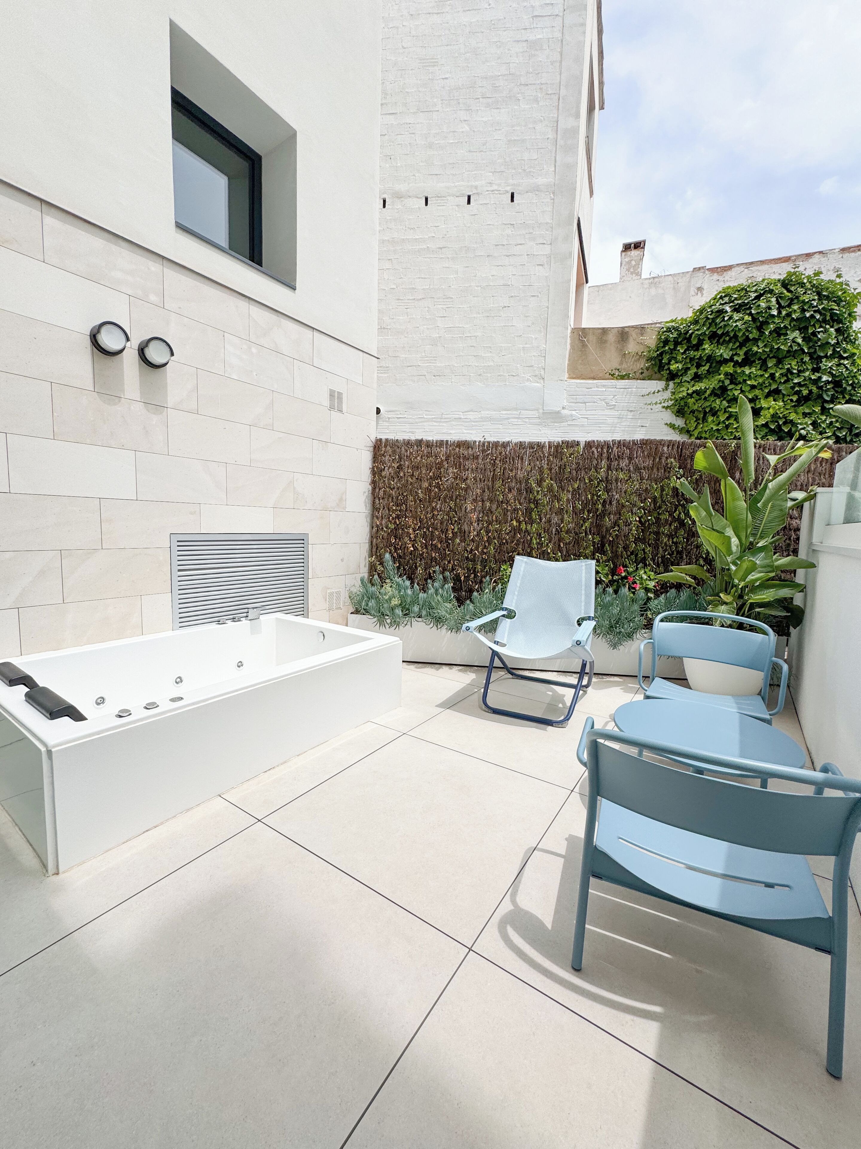 Un baño exterior moderno con una bañera grande, dos luces empotradas en la pared y una zona de estar con una mesa y sillas.