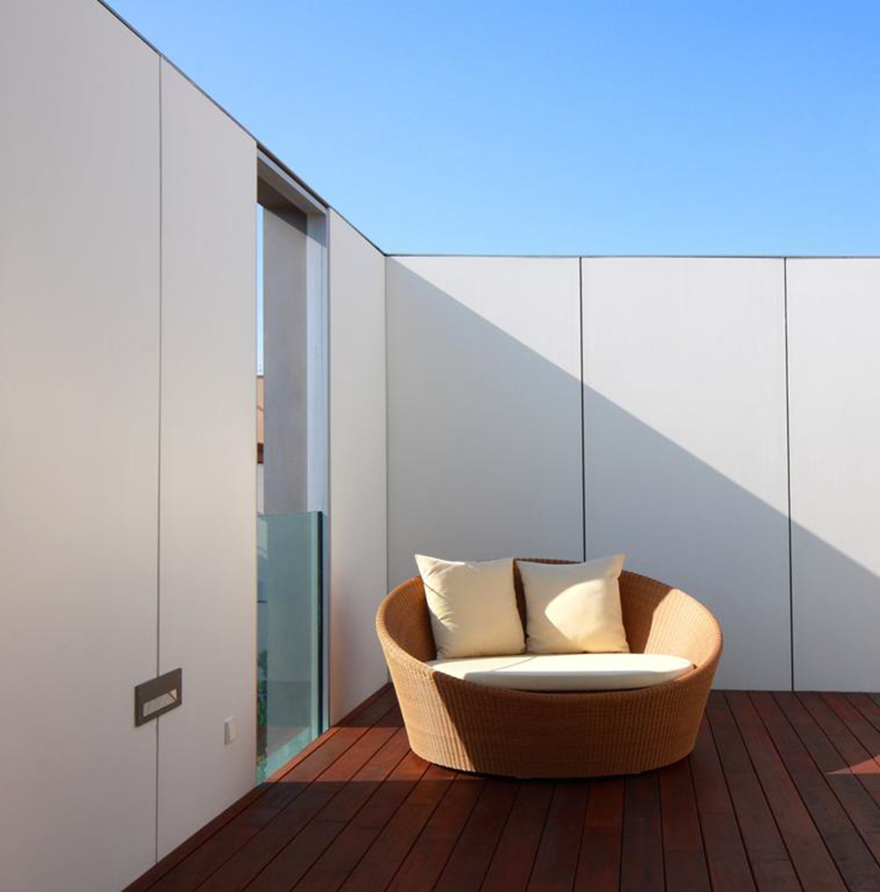 A modern outdoor seating area with a wicker chair and cushions, a wooden deck, and a glass door leading to an interior space.