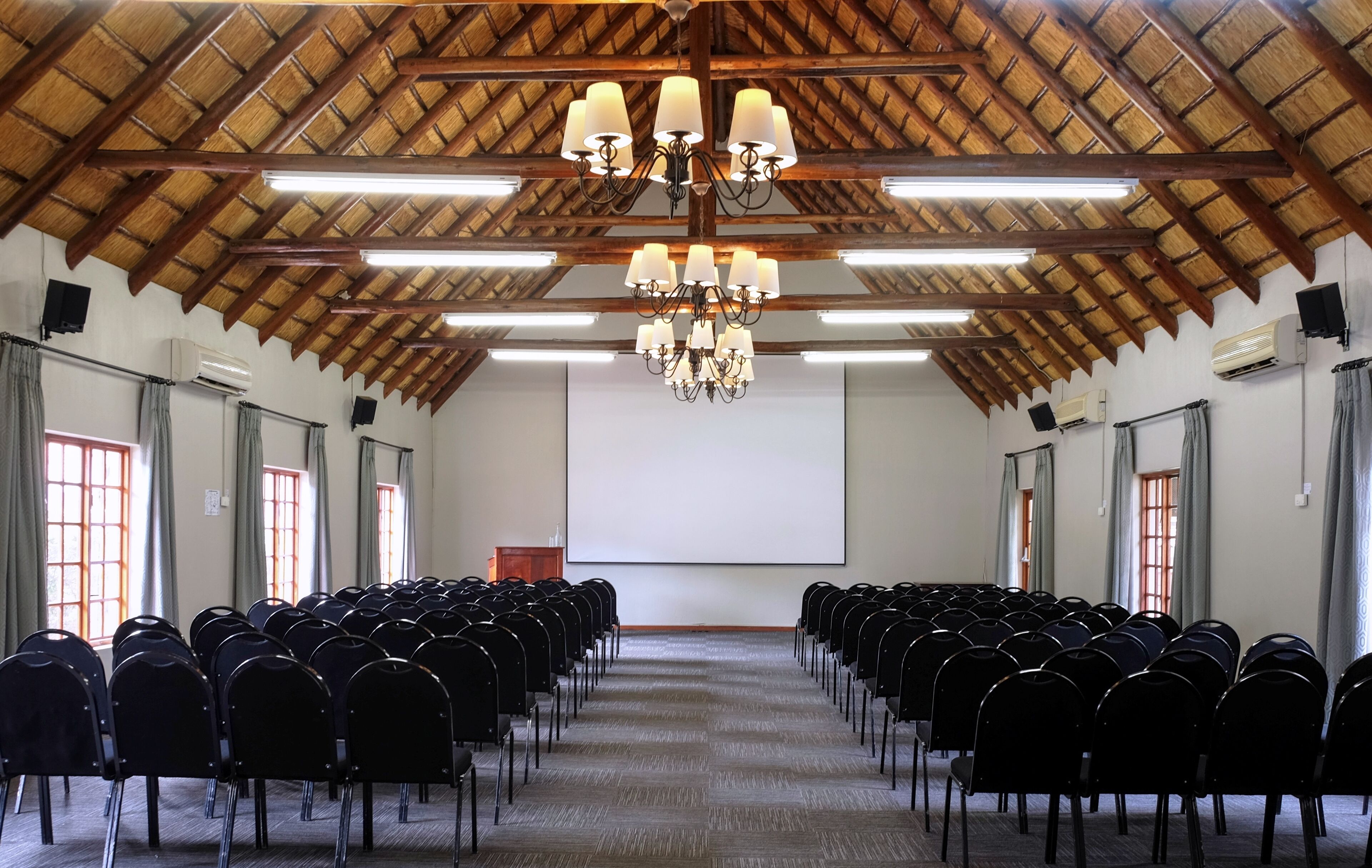 Un salón de conferencias con hileras de sillas negras, una pantalla de proyector y un techo de madera con candelabros.