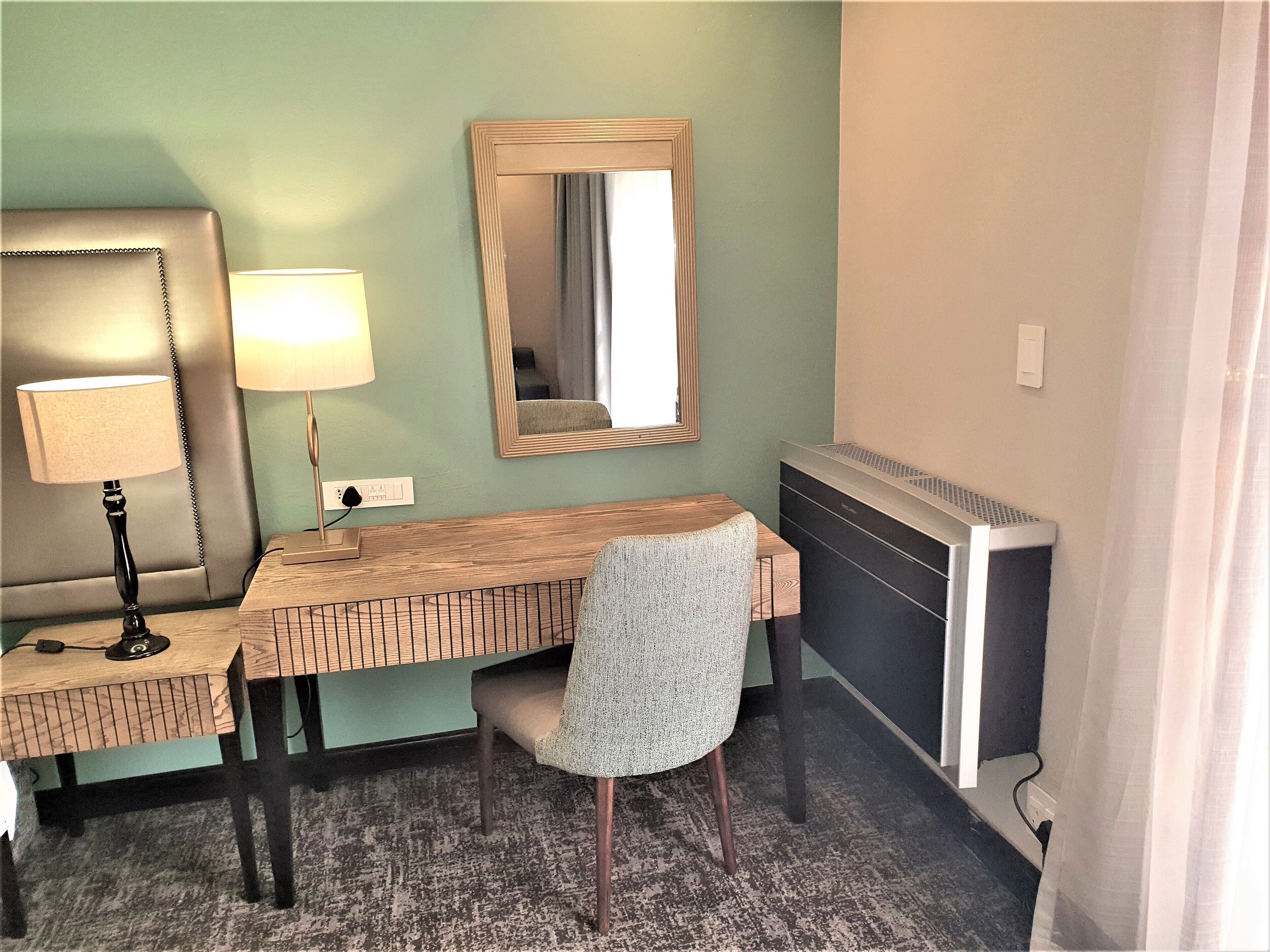 Habitació d'hotel amb escriptori de fusta, una cadira, una làmpada, un mirall i un radiador.