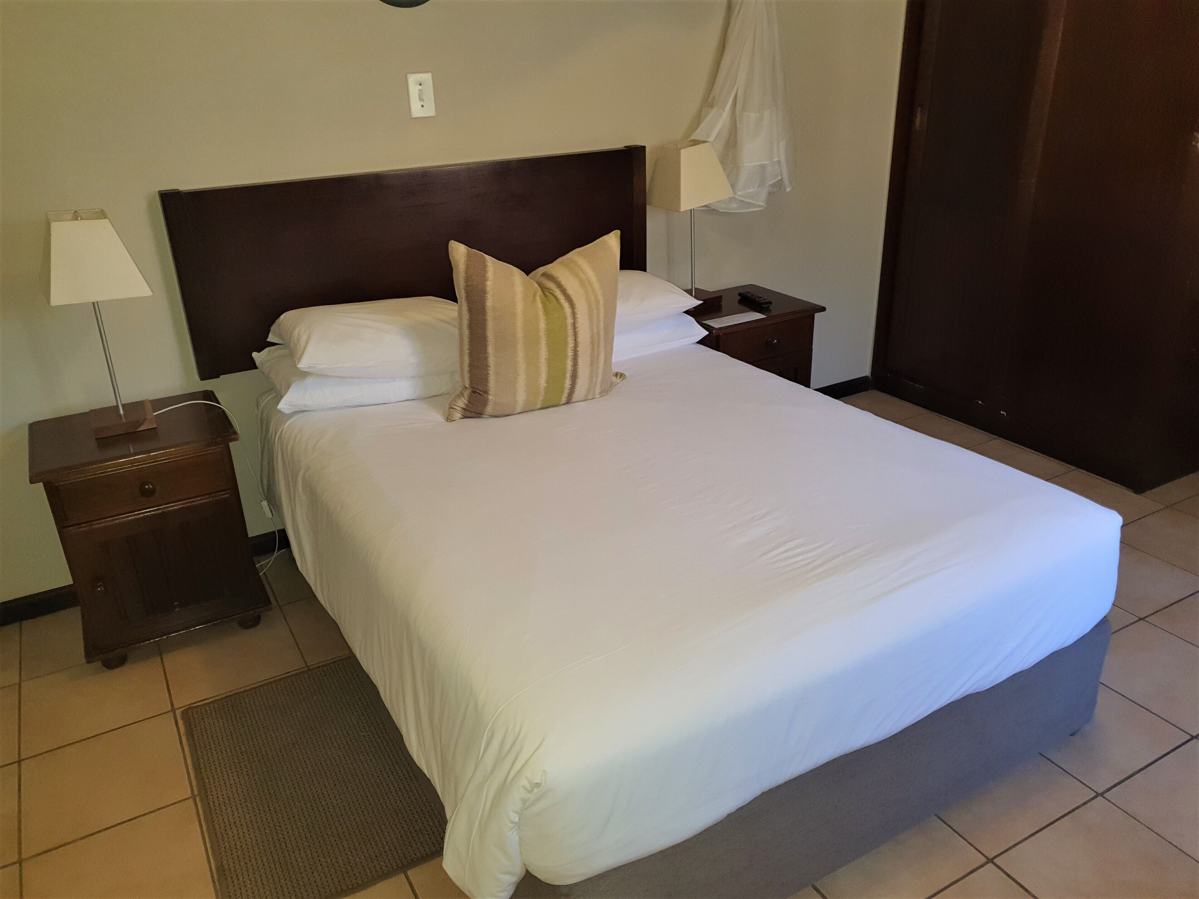 A neatly made bed with white linens and striped pillows, a bedside table with a lamp, and a small drawer.