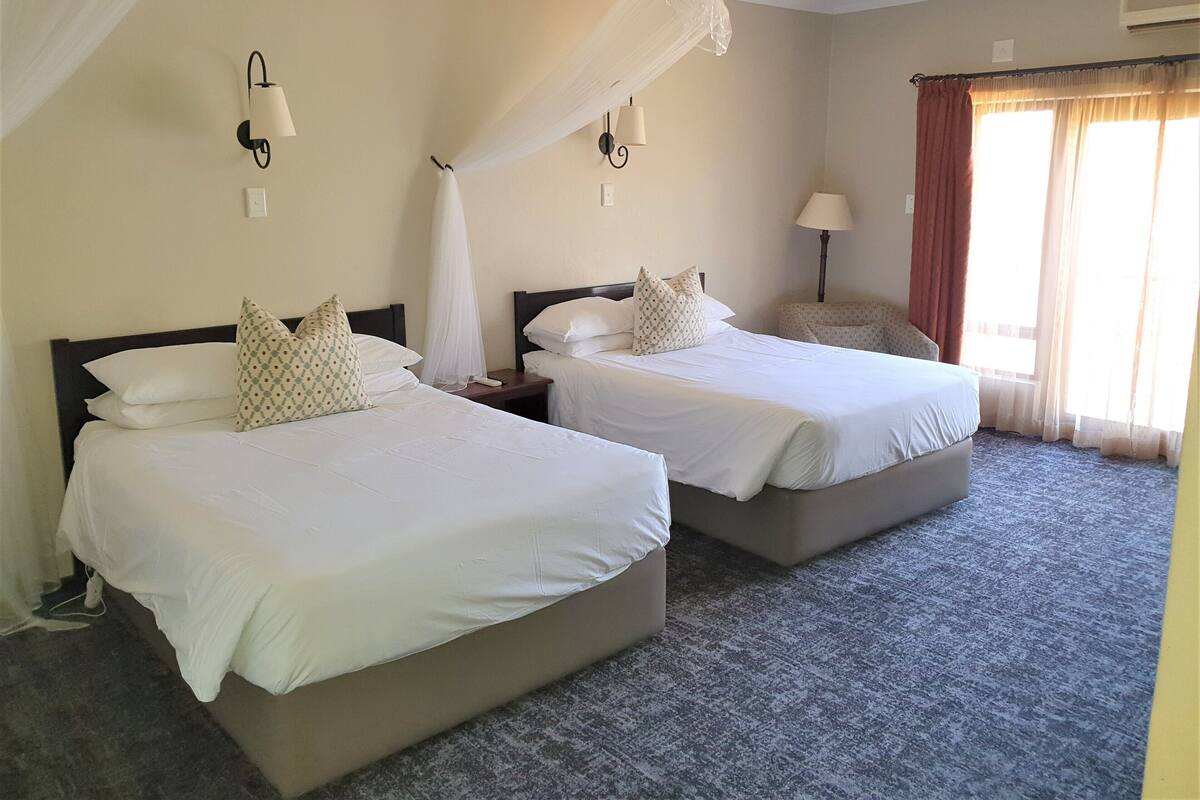 Habitación de hotel con dos camas, una mesita de noche, una lámpara y un ventanal con cortinas.