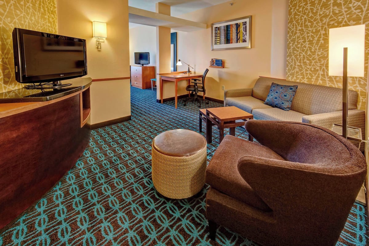 Une chambre d’hôtel équipée d’une télévision, d’un canapé, d’un fauteuil, d’une petite table et d’un bureau avec une lampe.