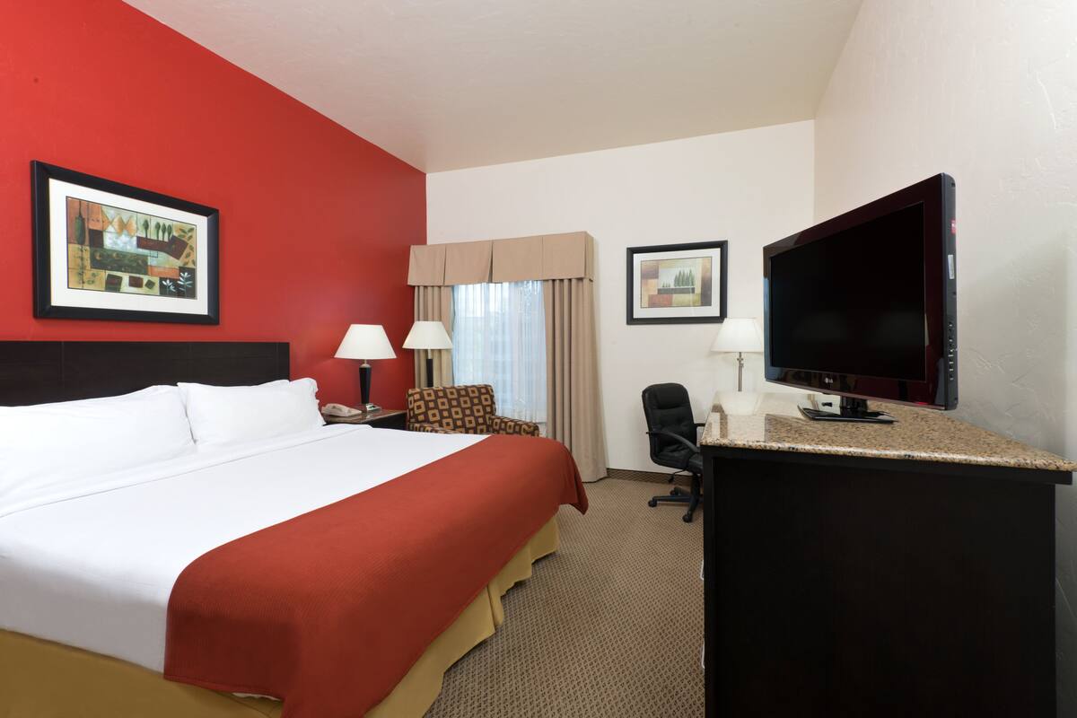 A hotel room with a large bed, a desk with a flat-screen TV, a chair, a lamp, and two framed pictures on the wall.