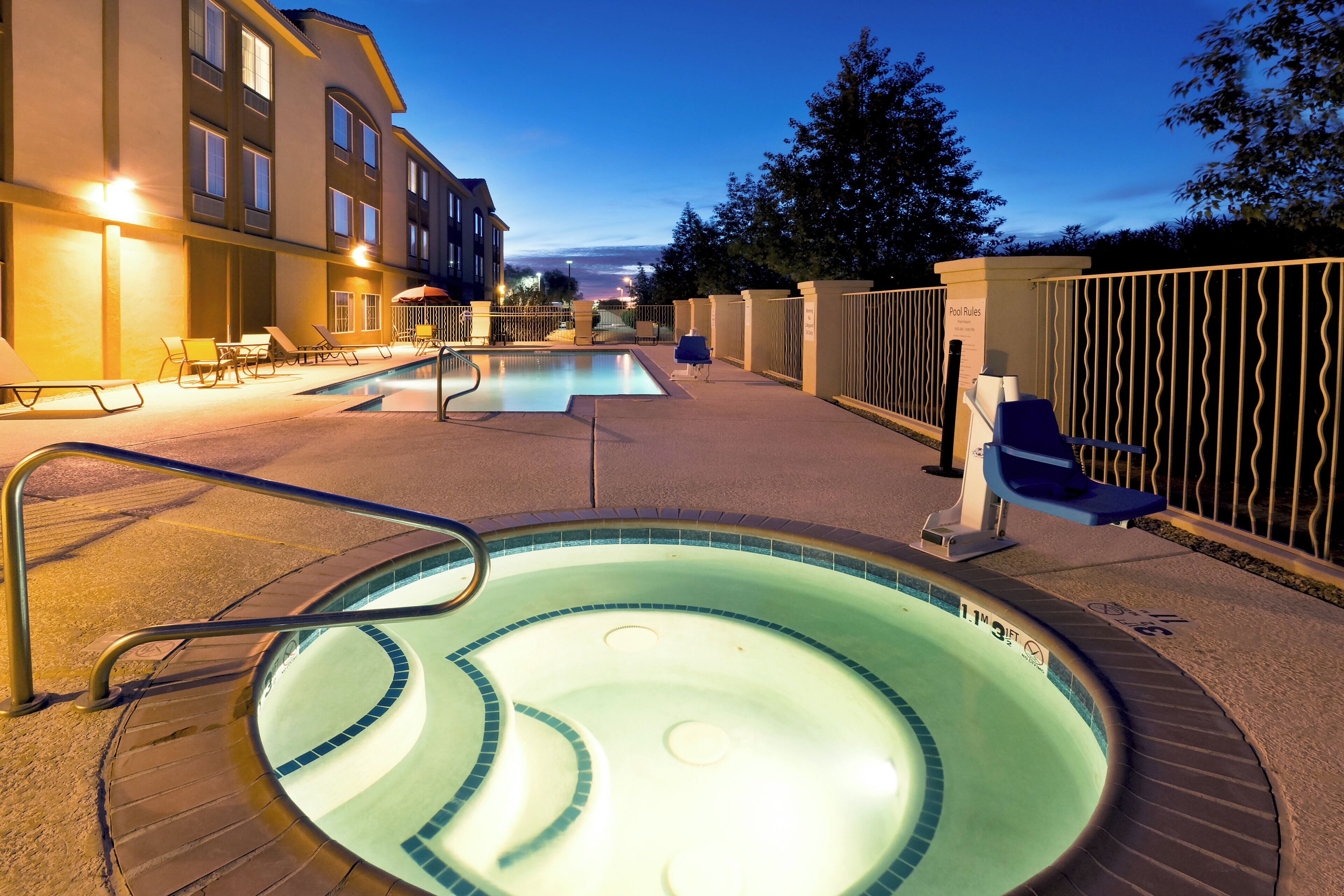 A swimming pool area with lounge chairs and a hot tub, surrounded by a fence and a building.