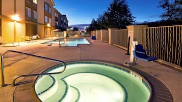 A swimming pool area with lounge chairs and a hot tub, surrounded by a fence and a building.