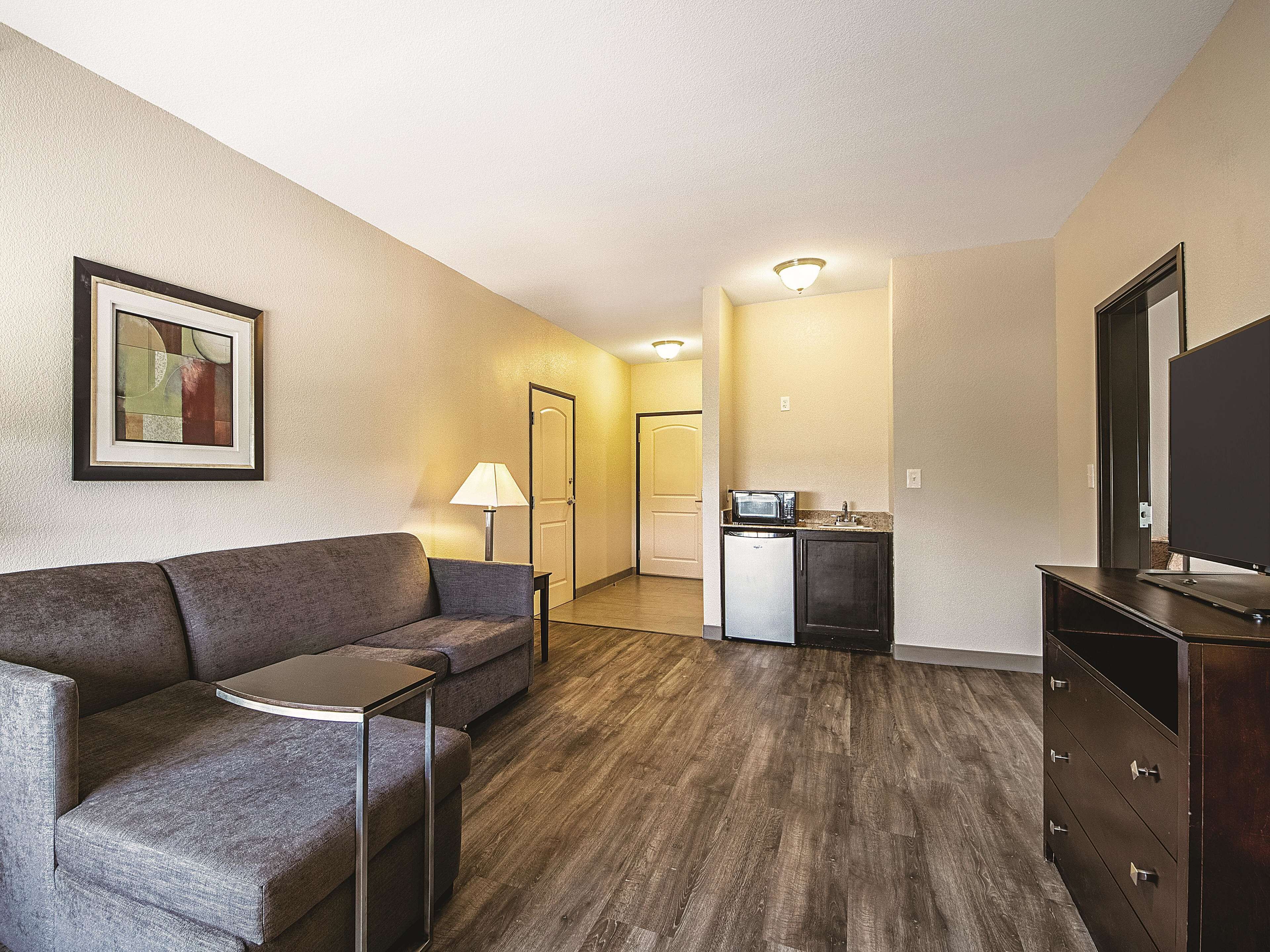 Une chambre d’hôtel compacte comprenant un canapé gris, une petite table, une kitchenette équipée d’un four à micro-ondes, une télévision et un placard.