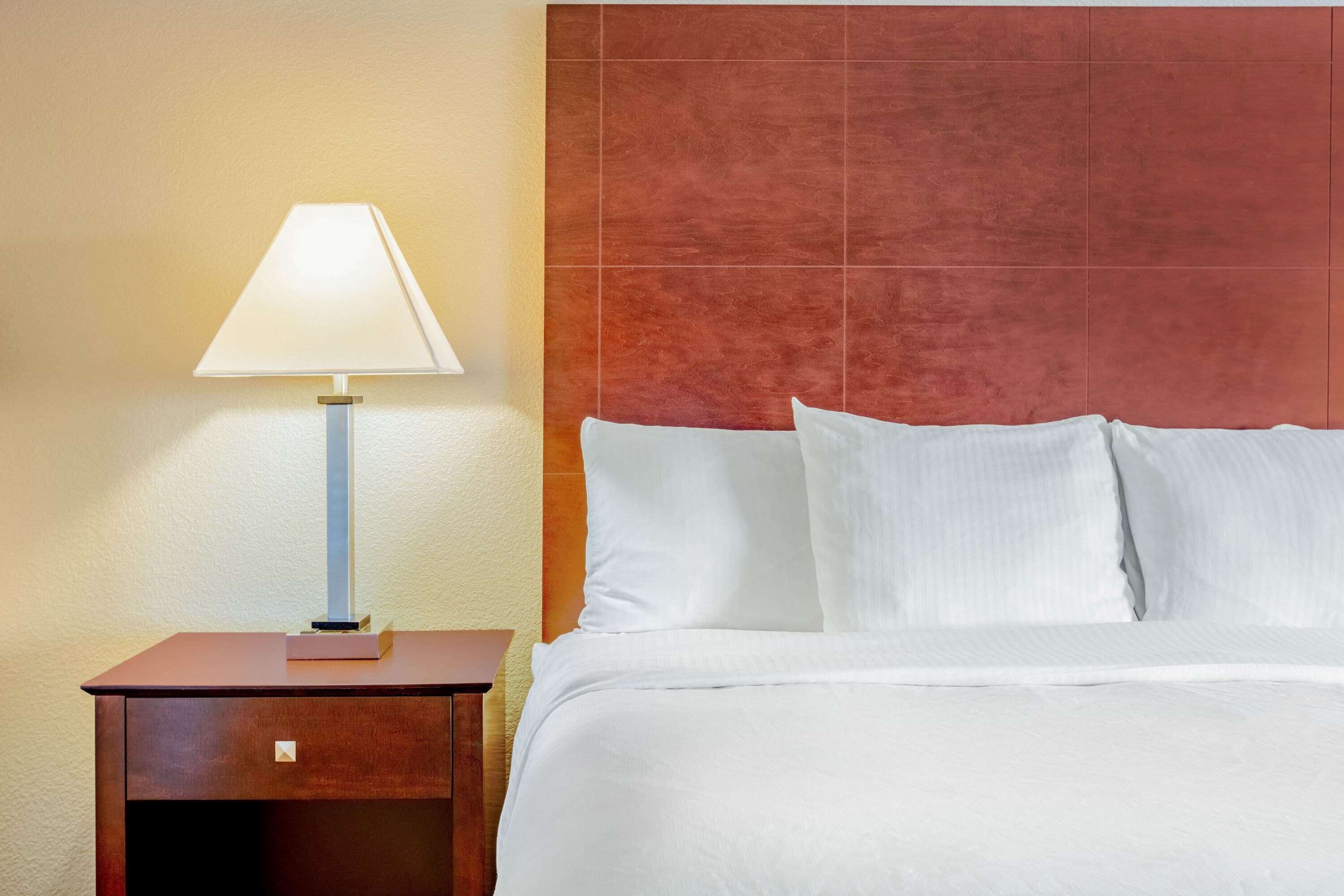 Una cama bien hecha con sábanas blancas, una mesita de noche de madera con una lámpara y un cabecero rojo texturizado.