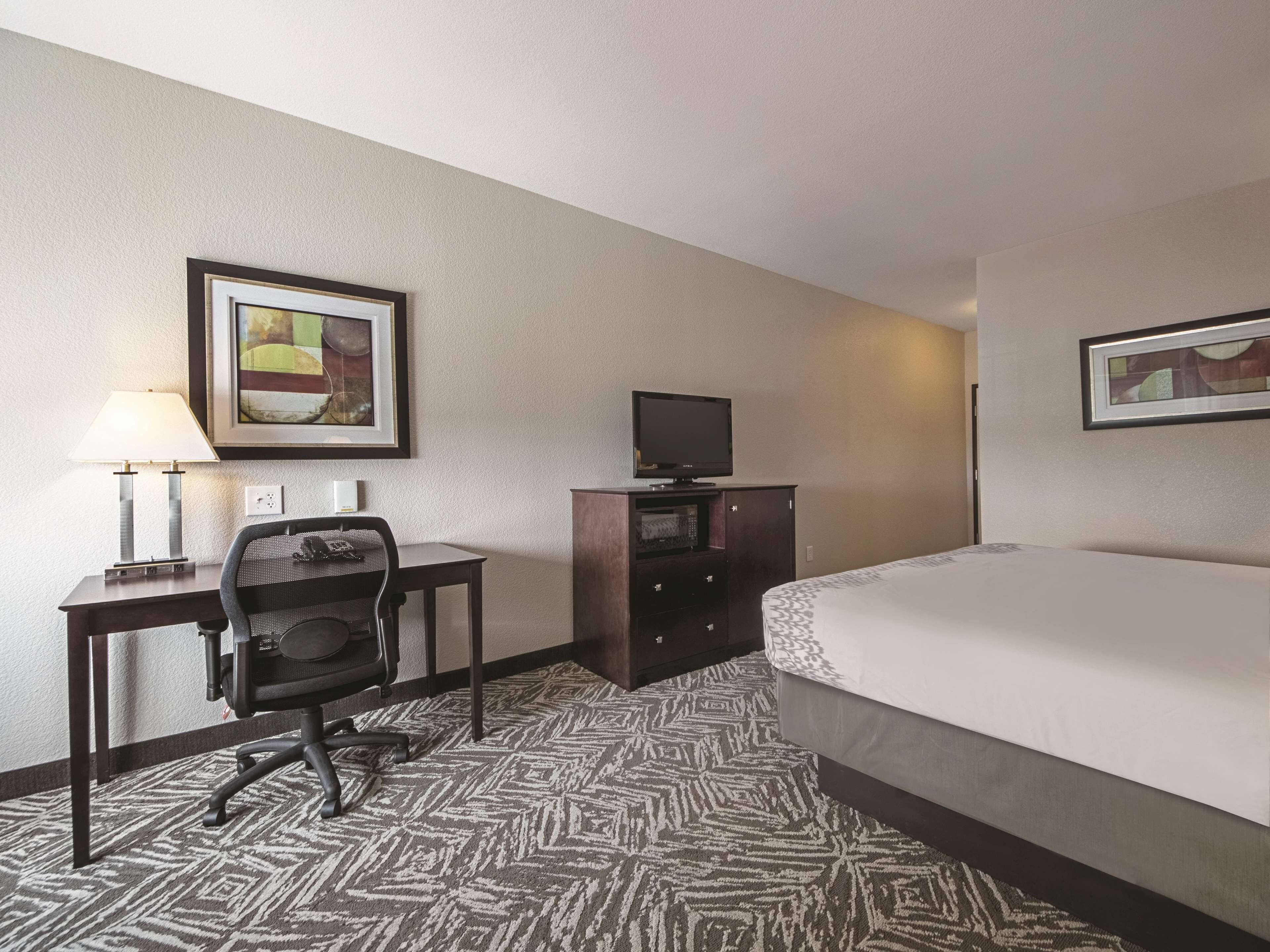 A hotel room with a bed, a desk with a chair, a television, and framed artwork on the walls.