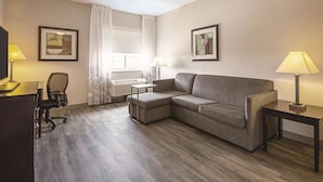 A hotel room with a grey sofa, a desk with a lamp, a chair, and two framed pictures on the wall. - La Quinta Inn & Suites by Wyndham McKinney (McKinney)