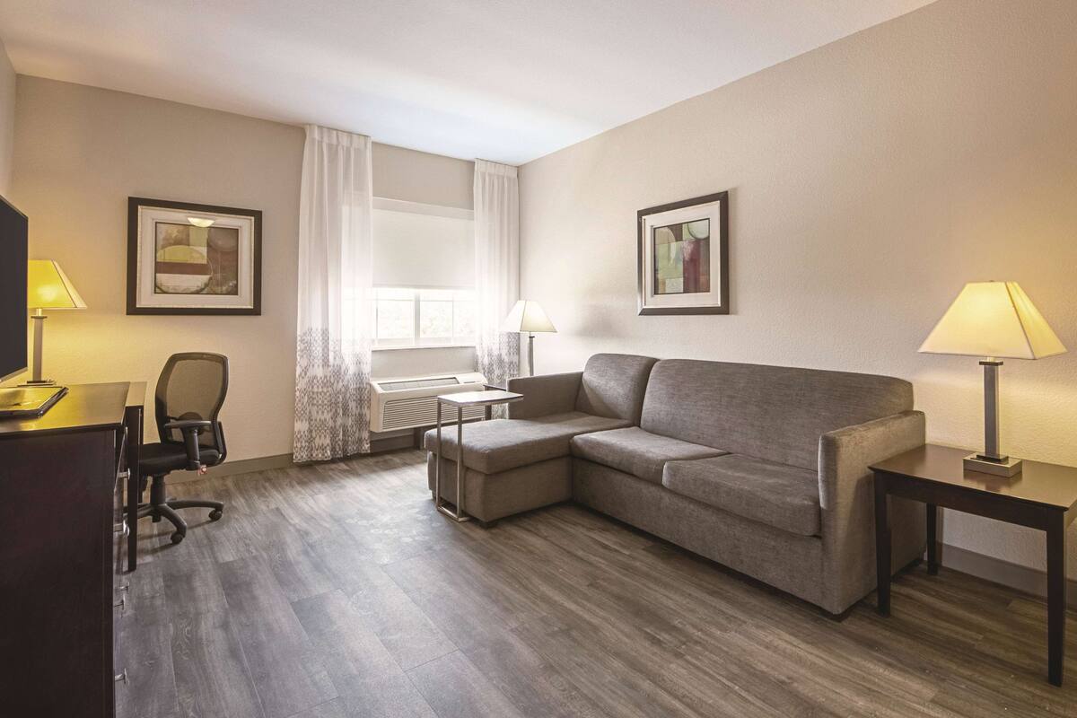 Ein Hotelzimmer mit einem grauen Sofa, einem Schreibtisch mit Lampe, einem Sessel und zwei gerahmten Bildern an der Wand.