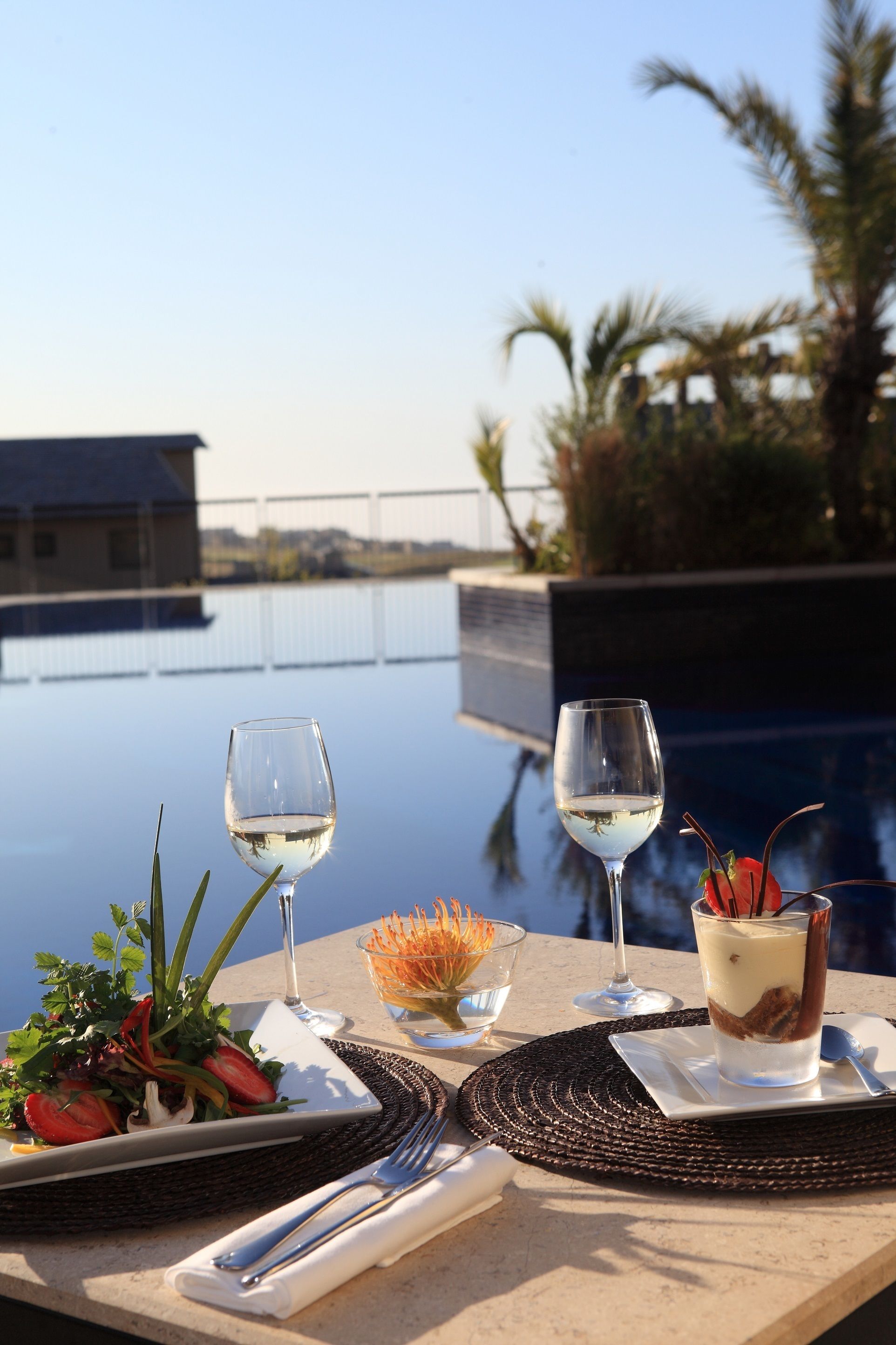 Una mesa de comedor con una copa de vino blanco, un postre y un plato de comida, con vistas a una piscina y palmeras.