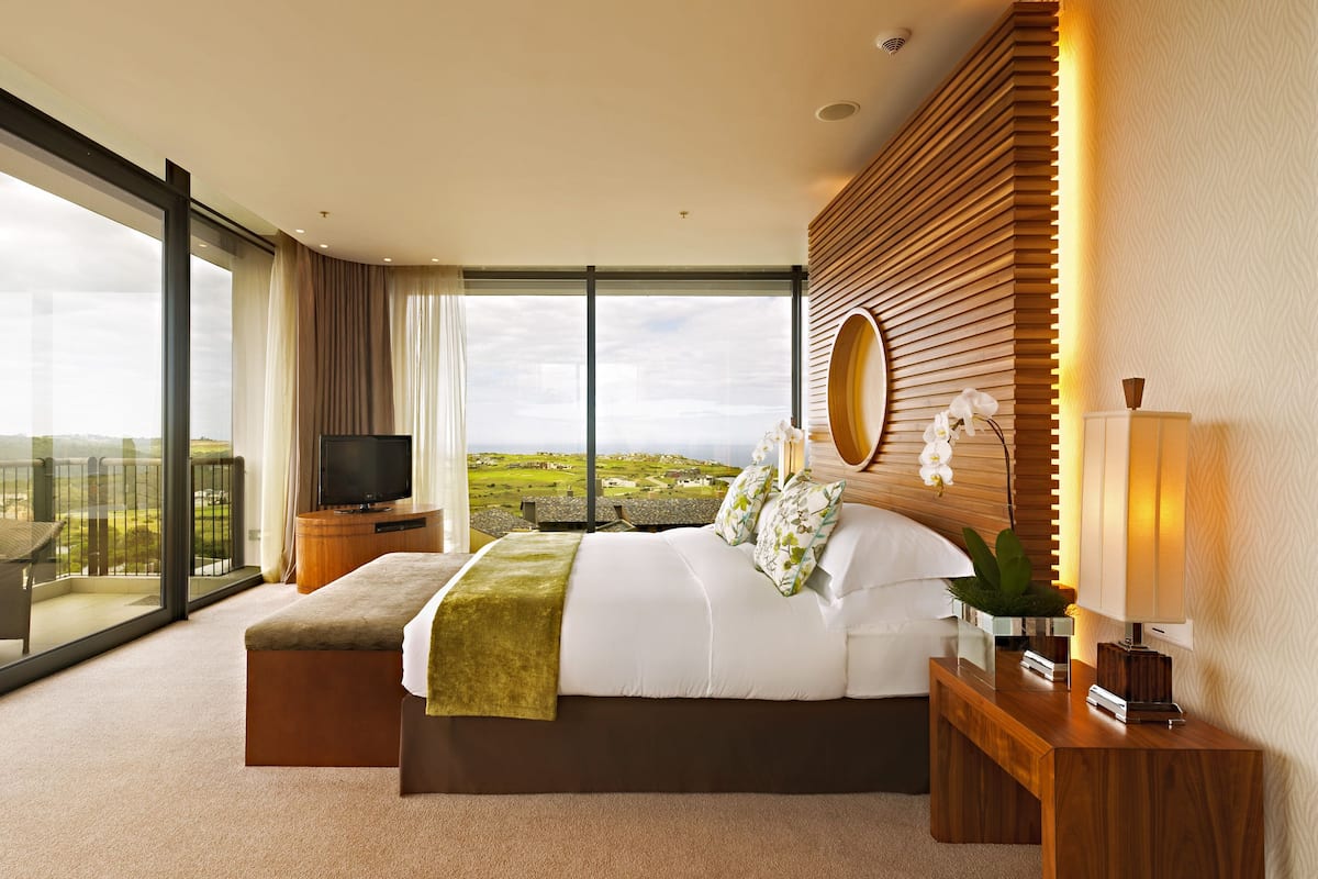 Une chambre d’hôtel moderne dotée d’un grand balcon, d’un lit agrémenté d’un couvre-lit vert, d’une télévision et d’une tête de lit en bois.