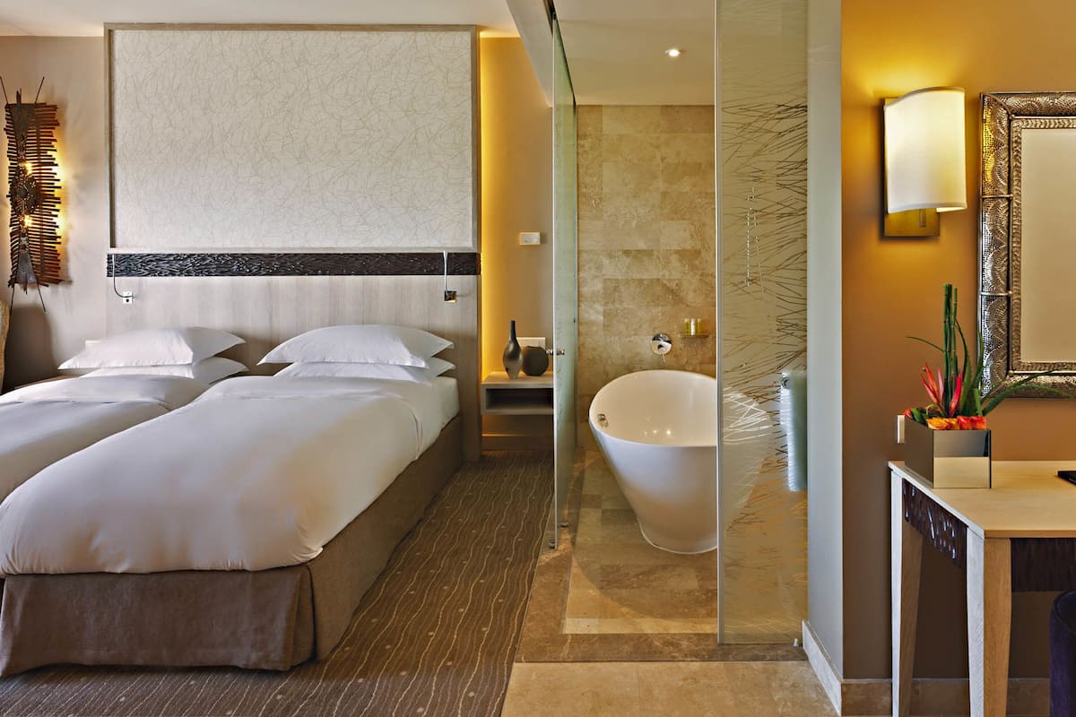 Une chambre d’hôtel avec deux lits, une grande baignoire, un bureau avec un miroir et un mur agrémenté d’un luminaire décoratif.