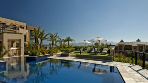 A swimming pool with lounge chairs and umbrellas, surrounded by palm trees and modern buildings. - Oubaai Hotel Golf & Spa (Herolds Bay)