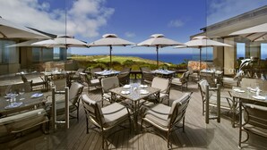 An outdoor dining area with tables and chairs, large umbrellas, and a view of the ocean. - Oubaai Hotel Golf & Spa (Herolds Bay)