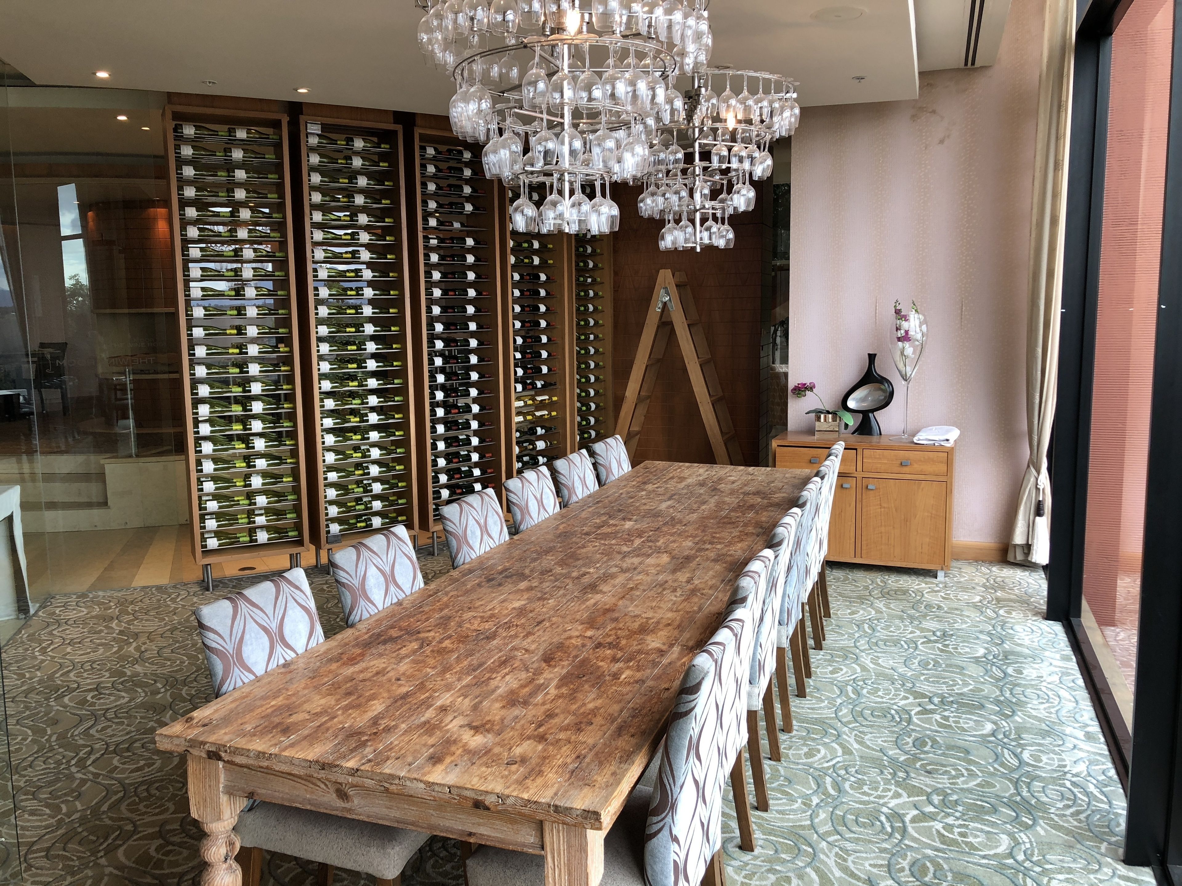 A long wooden table with patterned cushions, surrounded by tall wine racks, a wooden cabinet, and a decorative chandelier.