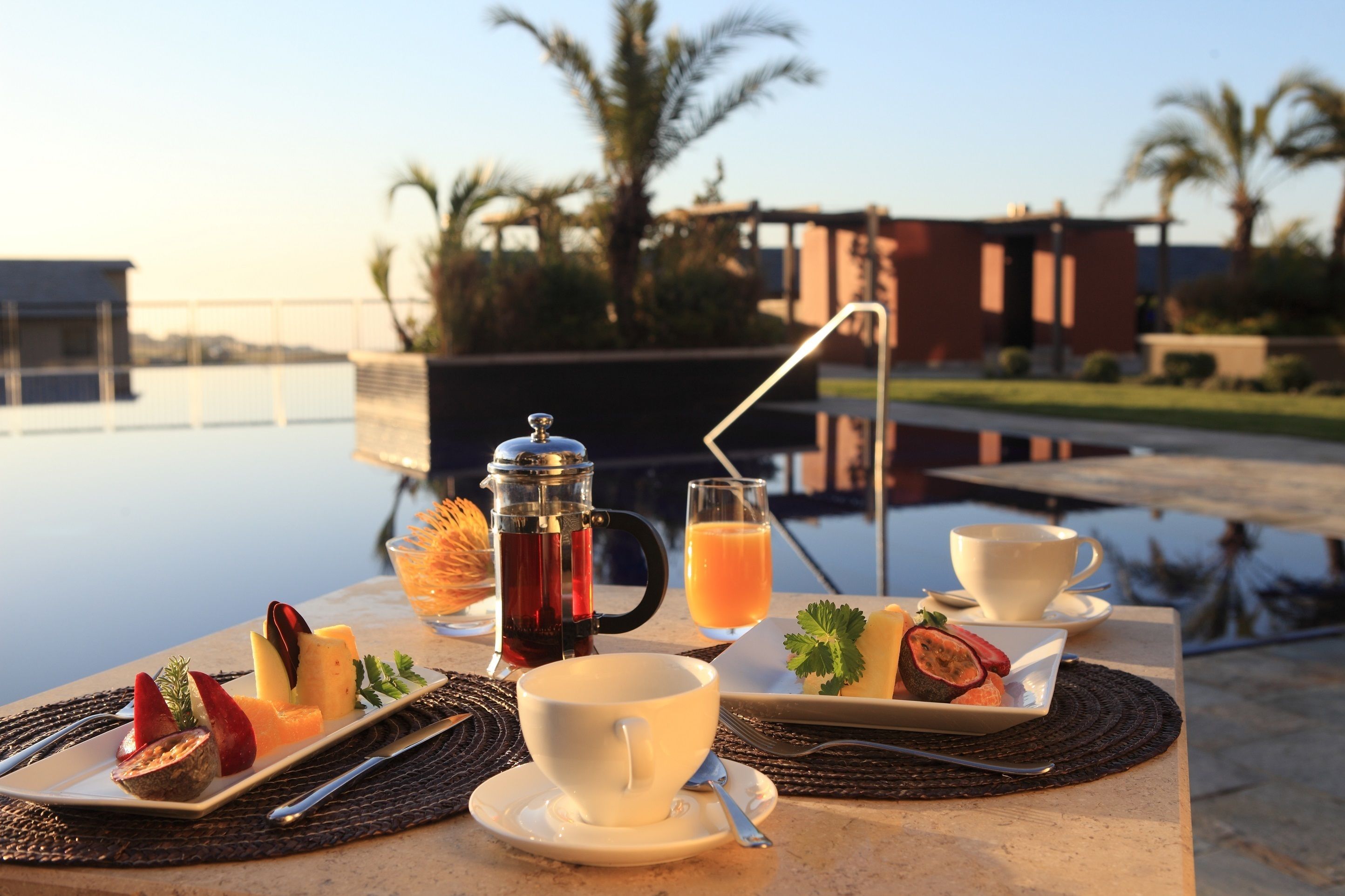 A breakfast setup with a coffee pot, juice, and fruit on a terrace with a view of palm trees and a building.