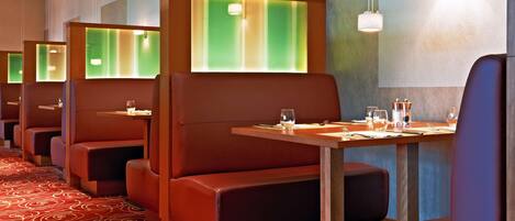 A row of booth-style seating with tables, each booth has a green-lit partition, and the floor has a red carpet with a spiral pattern.
