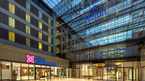 A modern hotel interior with glass walls, a visible Hilton Garden Inn sign, and a spacious lobby area.