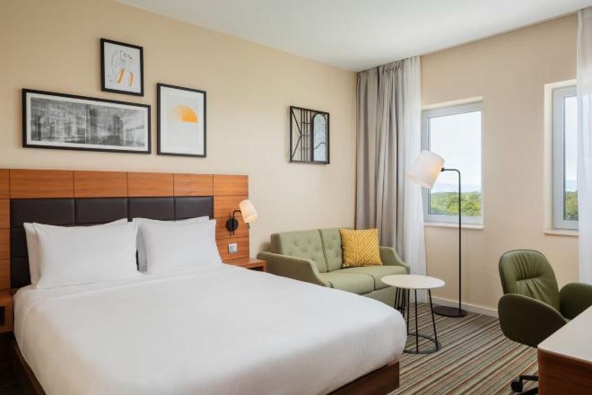 A hotel room with a large bed, a green sofa, a small round table, and a chair. There are framed pictures on the wall and a floor lamp.