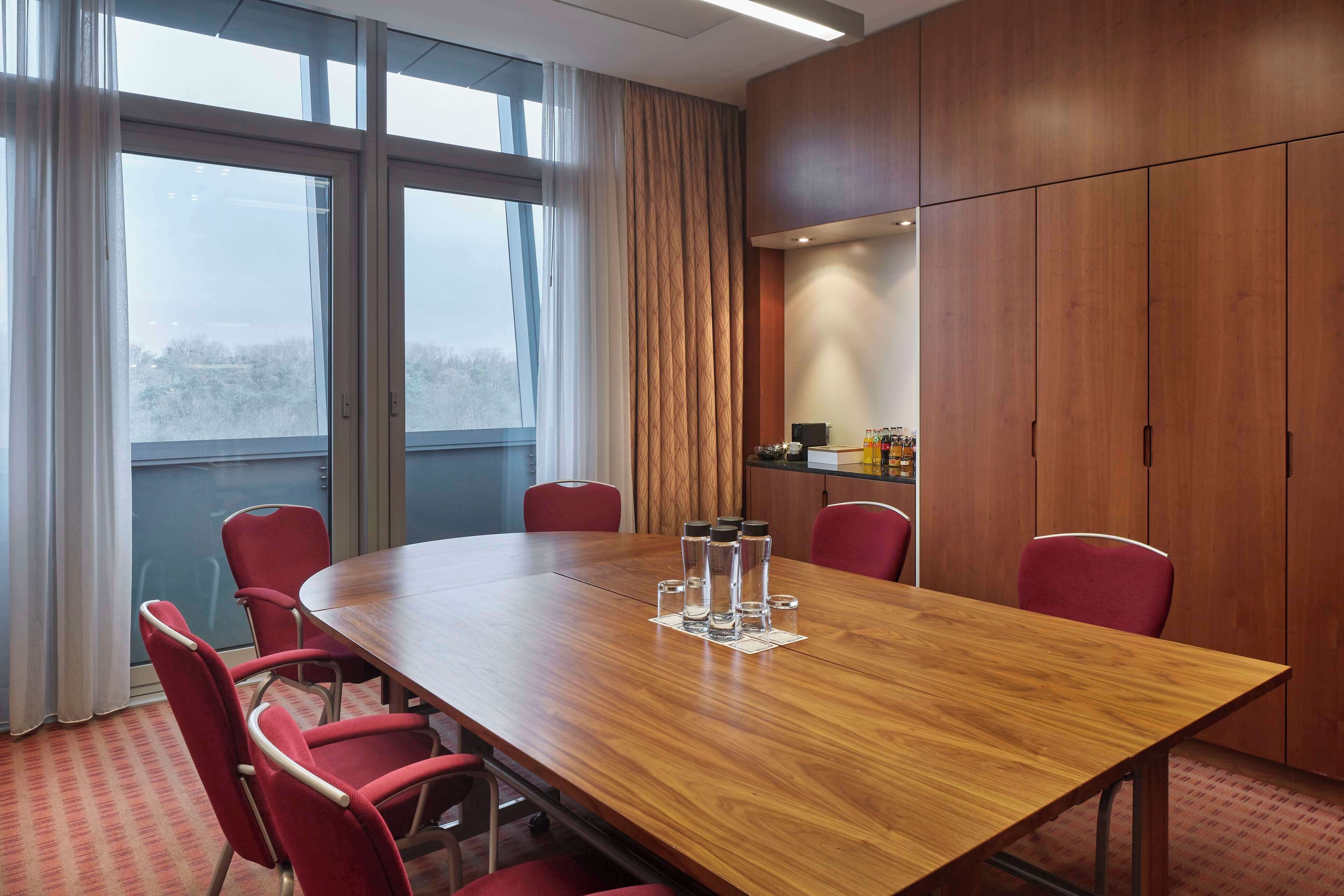 A conference room with a large wooden table, red chairs, and a view through large windows.
