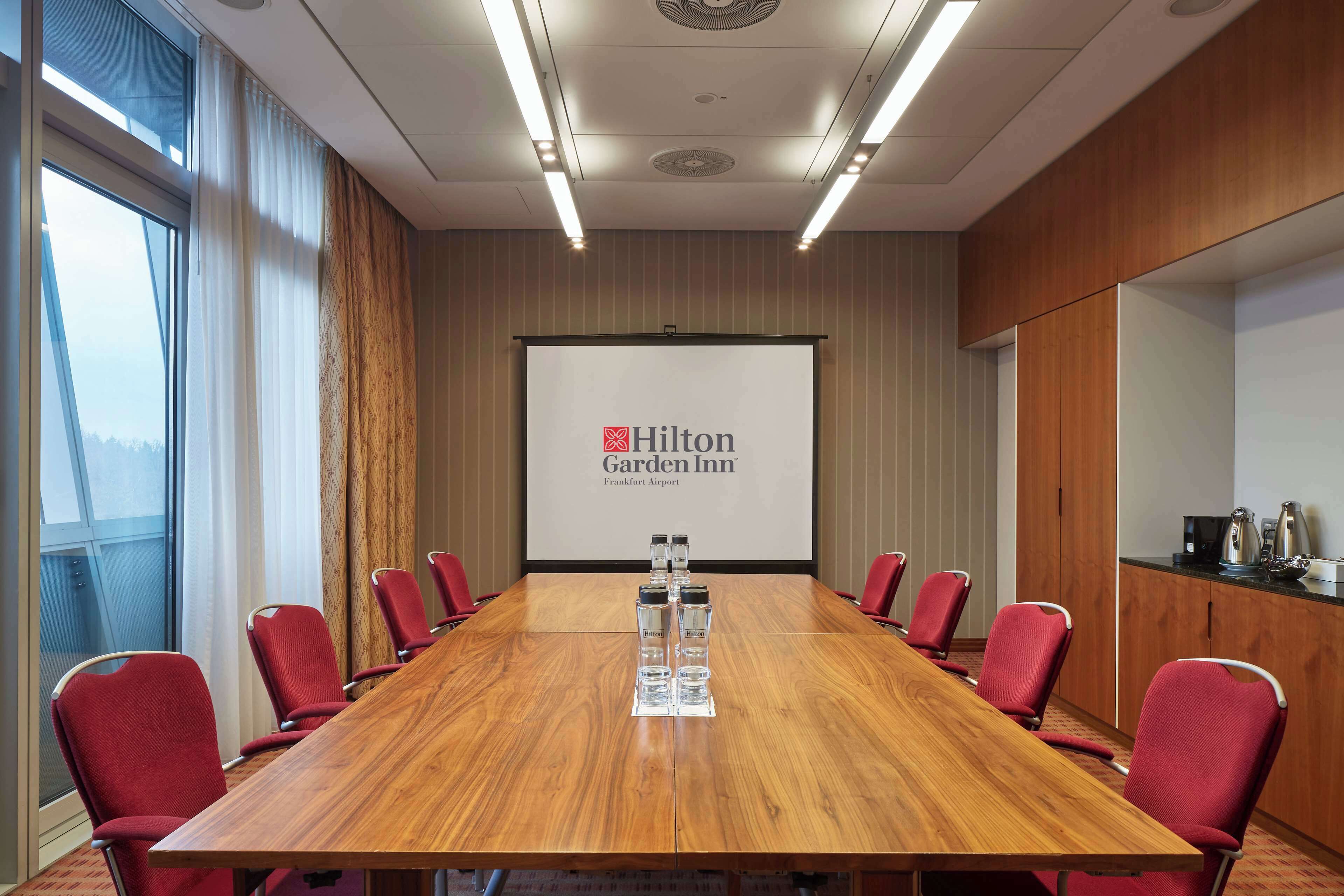 Ein Konferenzraum mit einem langen Holztisch, roten Stühlen, einer Projektionsleinwand mit dem Hilton Garden Inn Logo und Blick durch das Fenster.