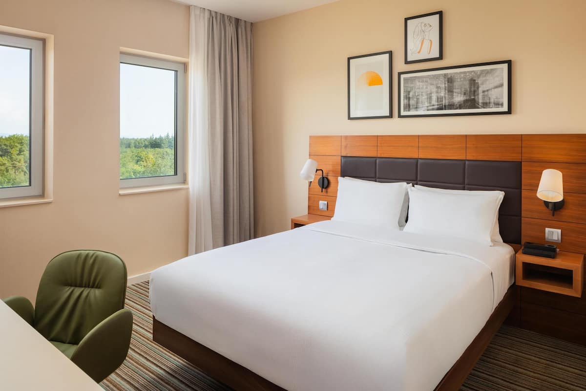 A hotel room with a large bed, two bedside tables, a chair, and two windows with curtains.