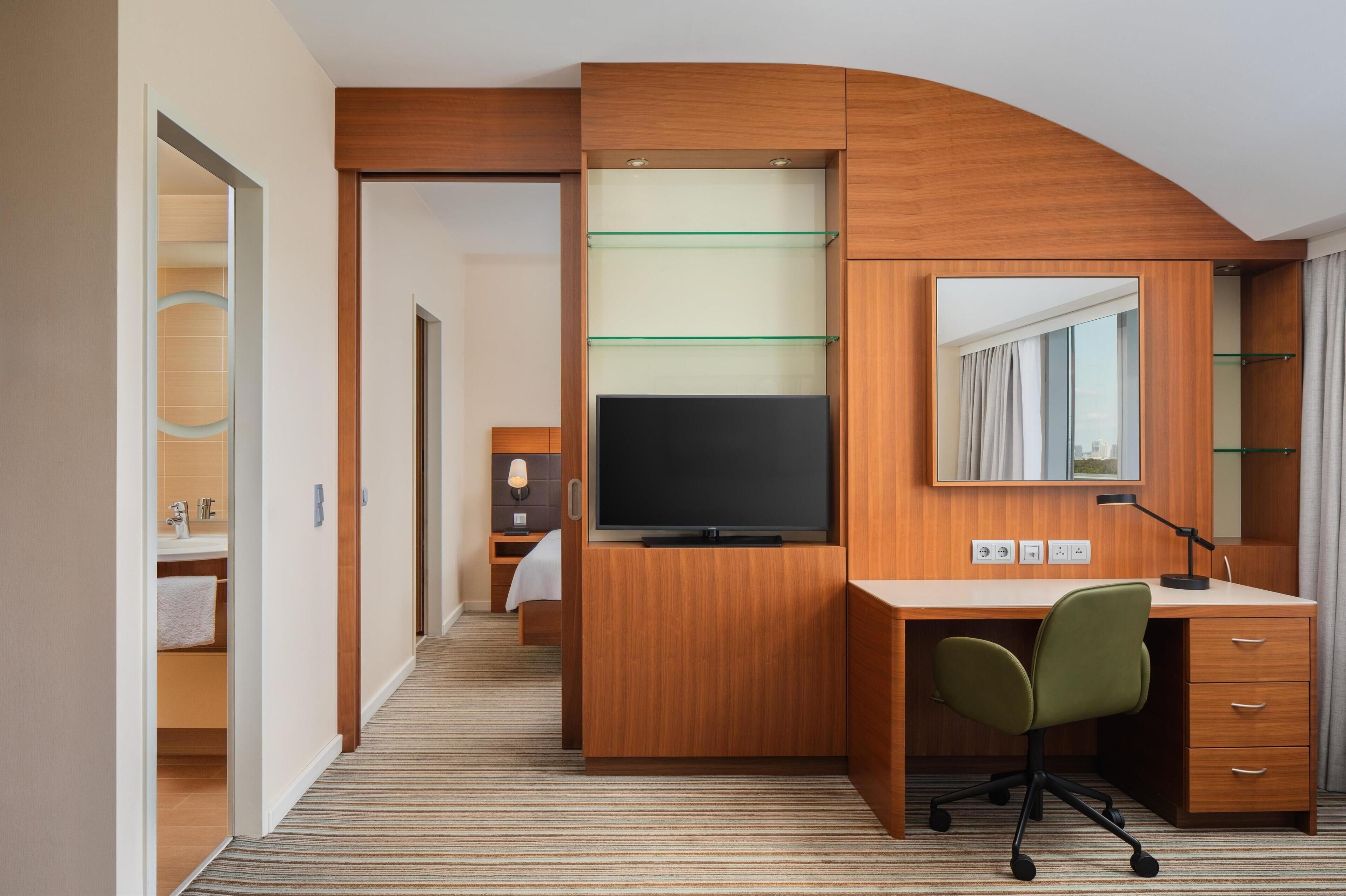 A hotel room with a wooden desk, a television, a mirror, and a glass shelf unit.