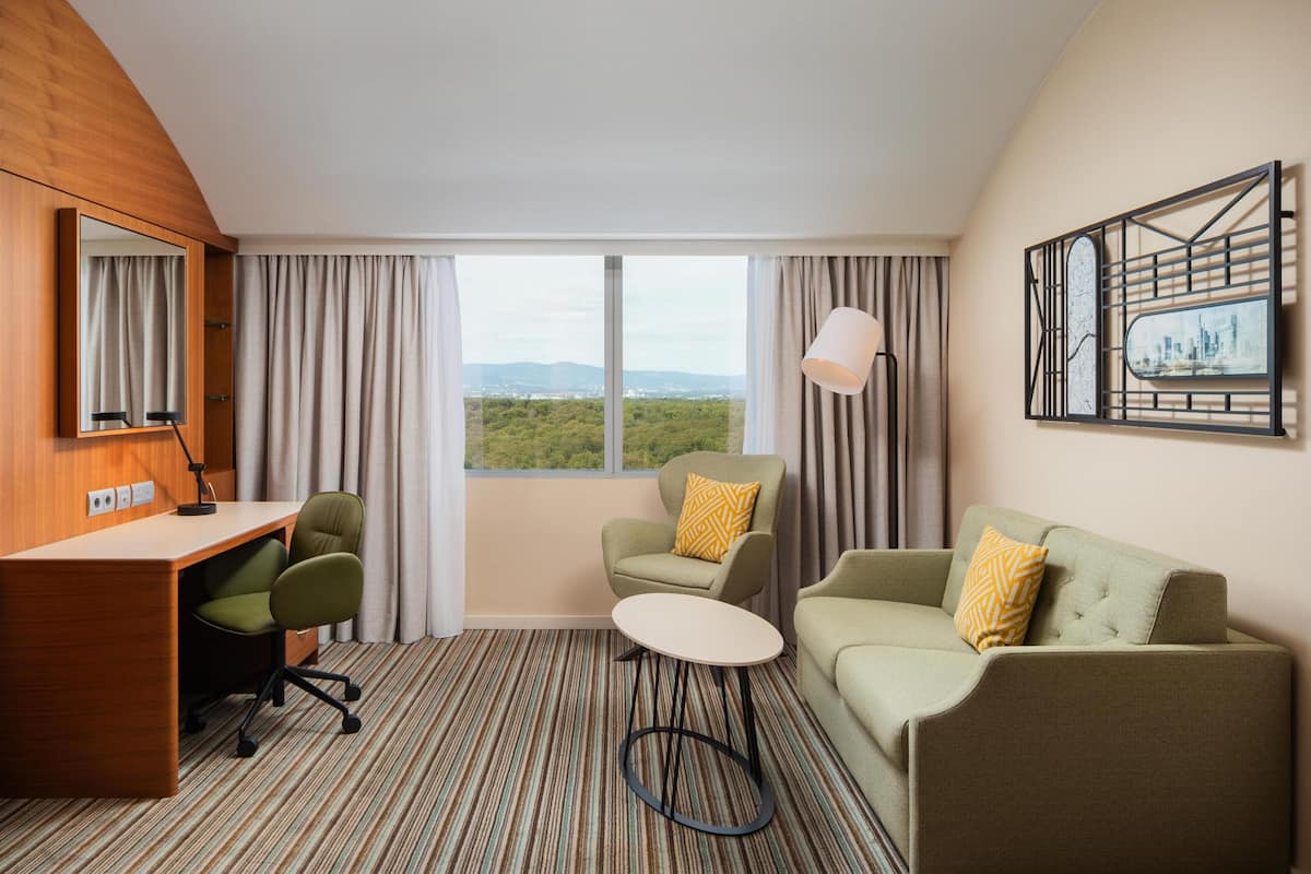 A hotel room with a desk, two chairs, a round table, a sofa, a large window with a view, and a wall-mounted artwork.
