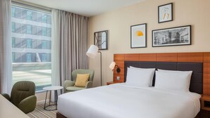 A hotel room with a large bed, a desk, a chair, and a view of a building through the window.