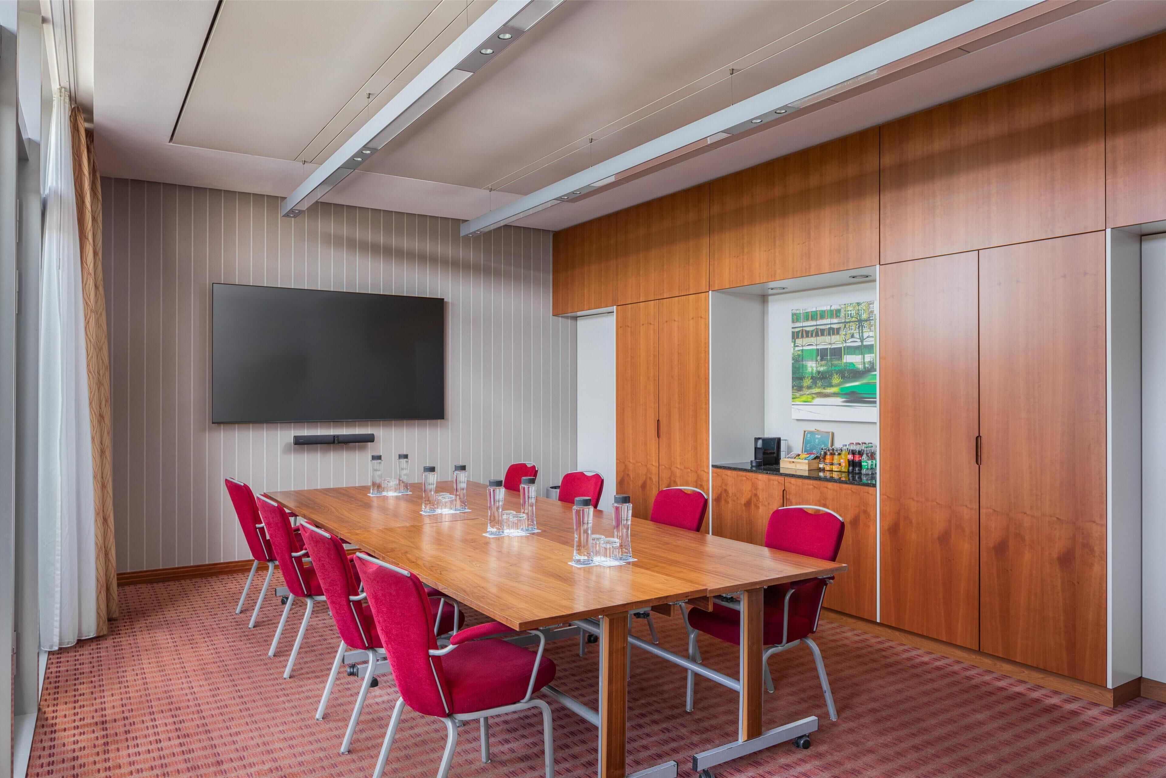A conference room with a long wooden table, red chairs, a flat-screen TV, and a view of a building through a window.