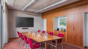 A conference room with a long wooden table, red chairs, a flat-screen TV, and a view of a building through a window.