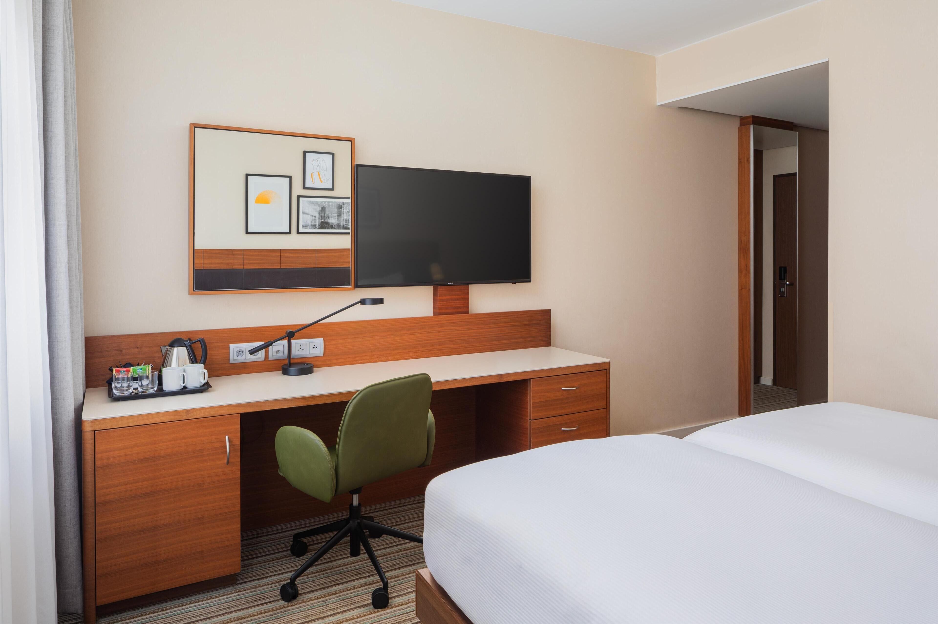 A hotel room with a wooden desk, a green office chair, a flat-screen TV, a bed, and a small seating area with a sofa.