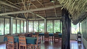 Un comedor con mesas y sillas de madera, ventanas amplias y un techo de paja.