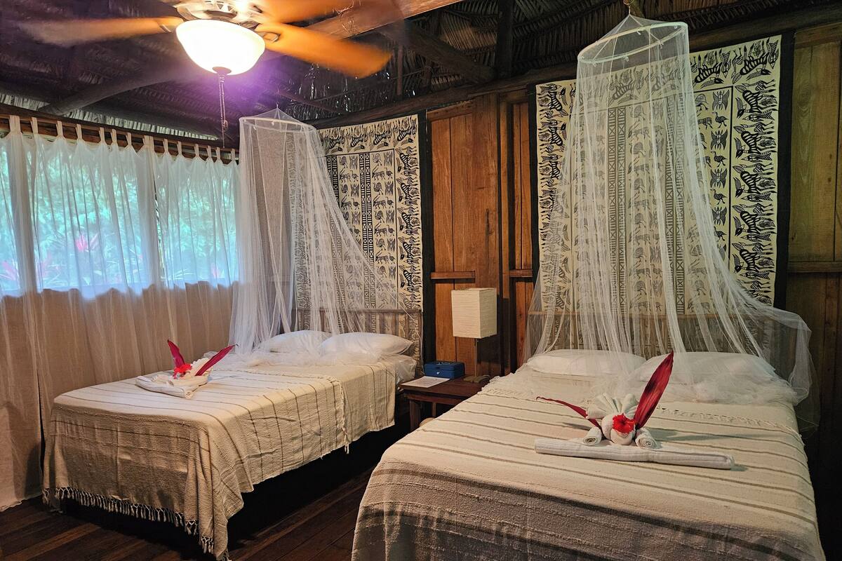 A room with two beds, each with a canopy and white bedding. There is a wooden floor and a ceiling fan.