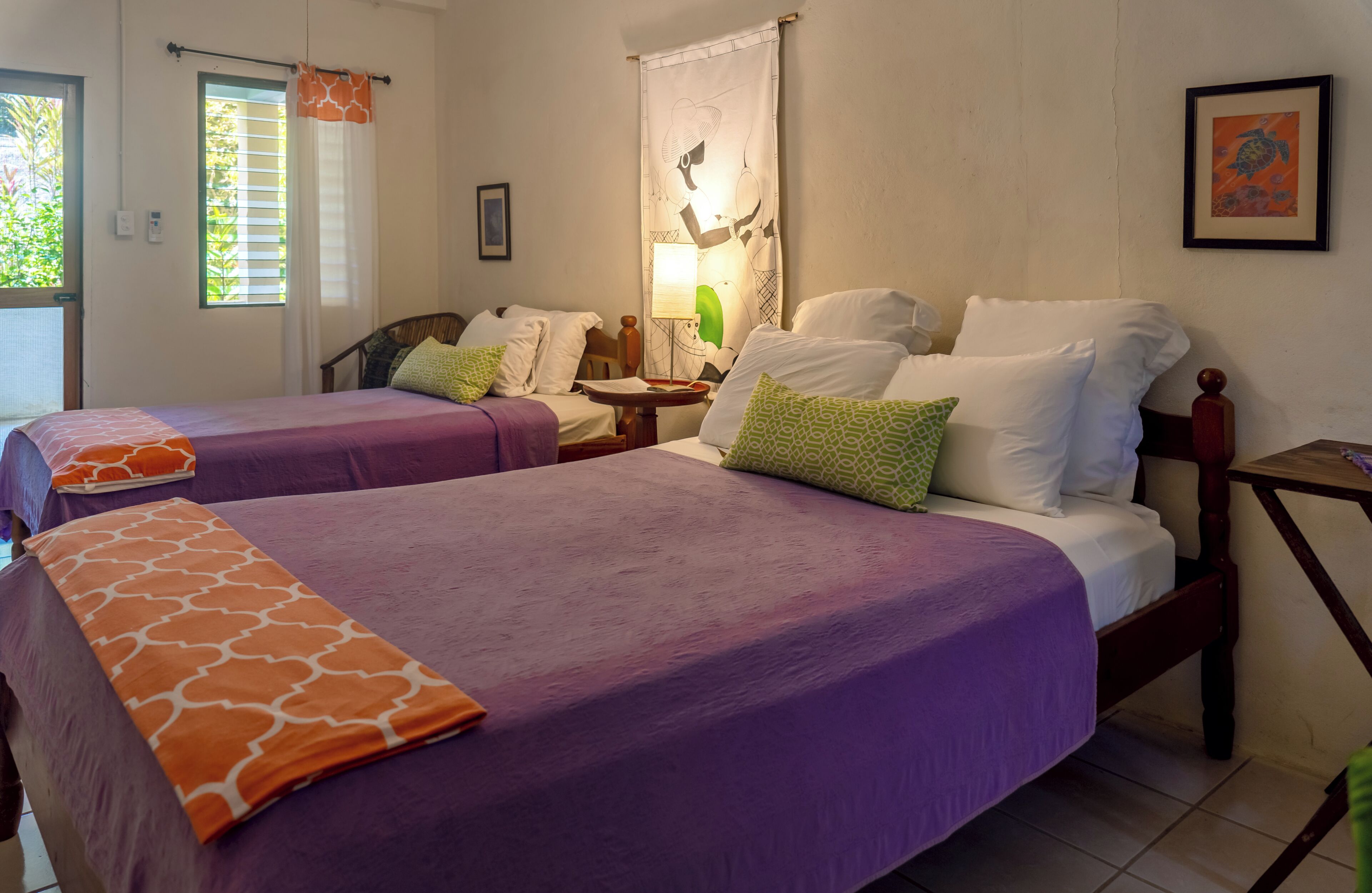 Une chambre d’hôtel avec deux lits, chacun recouvert d’une couvre-lit violette et d’un plaid orange à motifs. Il y a une fenêtre avec des stores, une lampe sur une table de chevet et un tableau encadré au mur.