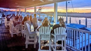 Un espace extérieur aménagé avec des tables et des chaises blanches, protégé par un auvent, et offrant une vue sur un port de plaisance rempli de bateaux.