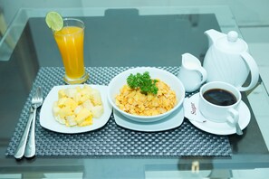 Frühstücksszene mit einer Schüssel Ananasstückchen, einer Schüssel Müsli mit Petersilie, einem Glas Orangensaft, einer Kaffeetasse und einem weißen Teekännchen auf einem Tisch mit Untersetzer aus Braidmaterial.